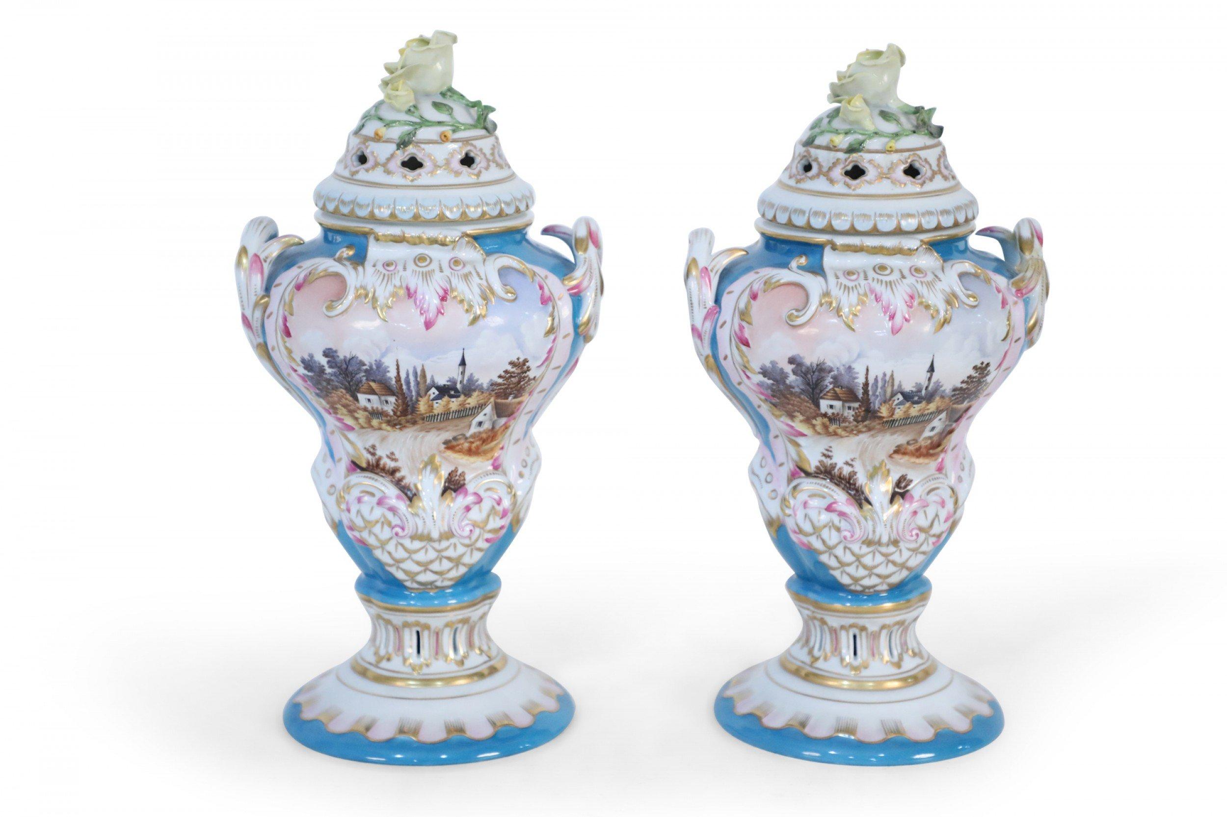 Pair of Hungarian 20th century porcelain urns with bright blue backgrounds and painted central pastoral scenes with gilt and pink details and lids with decorative cutouts topped with yellow porcelain roses (Marked, HEREND HUNGARY).