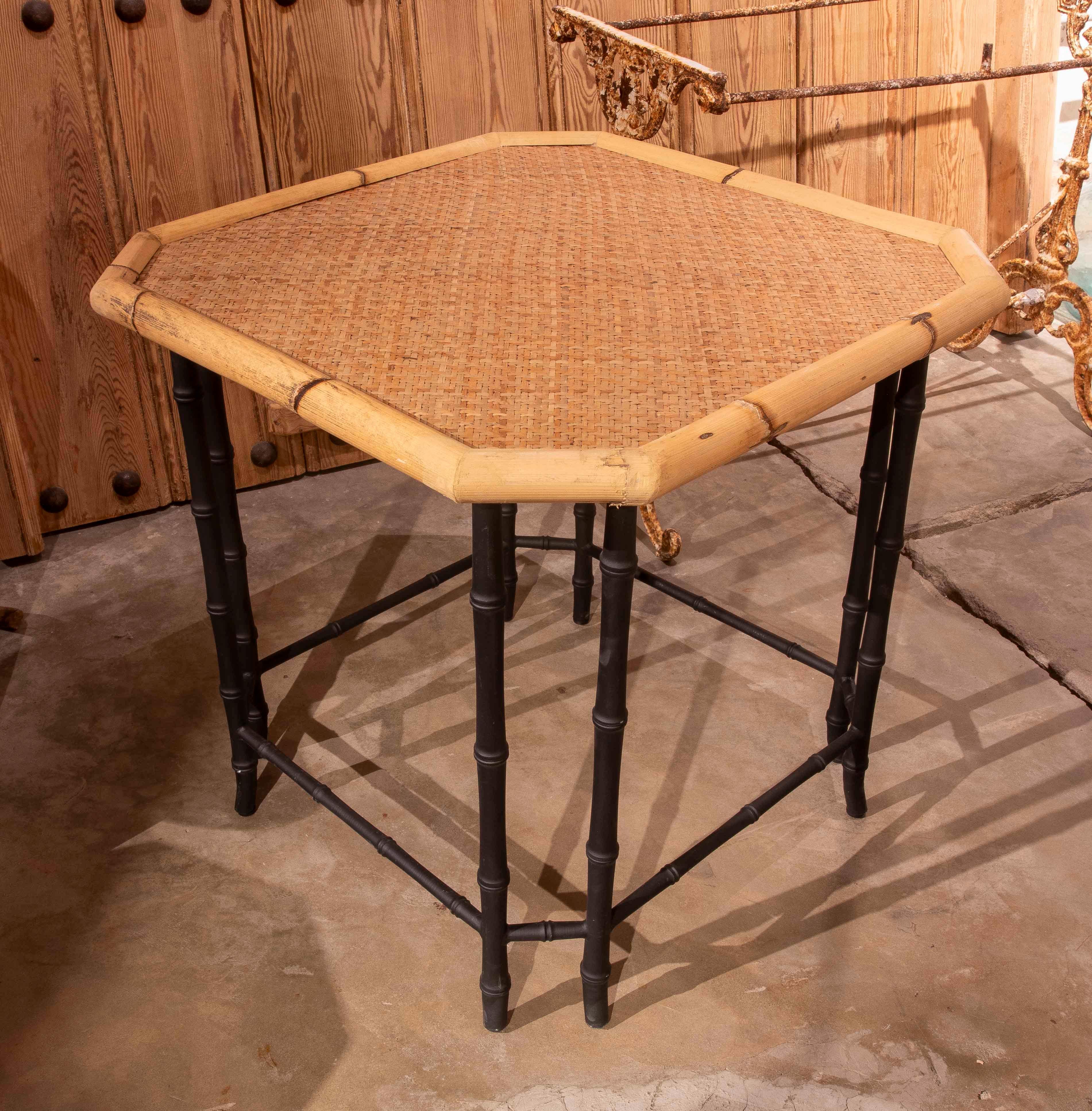Charming hexagonal side table, crafted with a natural bamboo frame and hand-woven rattan top, provides a warm, handcrafted feel. Its structure rests on black-painted metal legs with a bamboo-effect design, an elegant detail that enhances the piece's