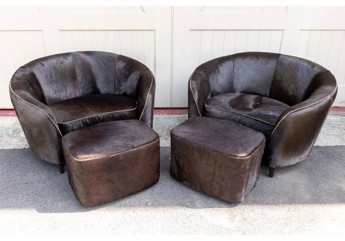 Large scale comfortable Club chairs with curved seats upholstered in black-brown cowhide with contrasting tan suede cord trim. Raised on cylindrical tapering front legs. With matching ottomans, without the cord trim but shaped to fit the curved