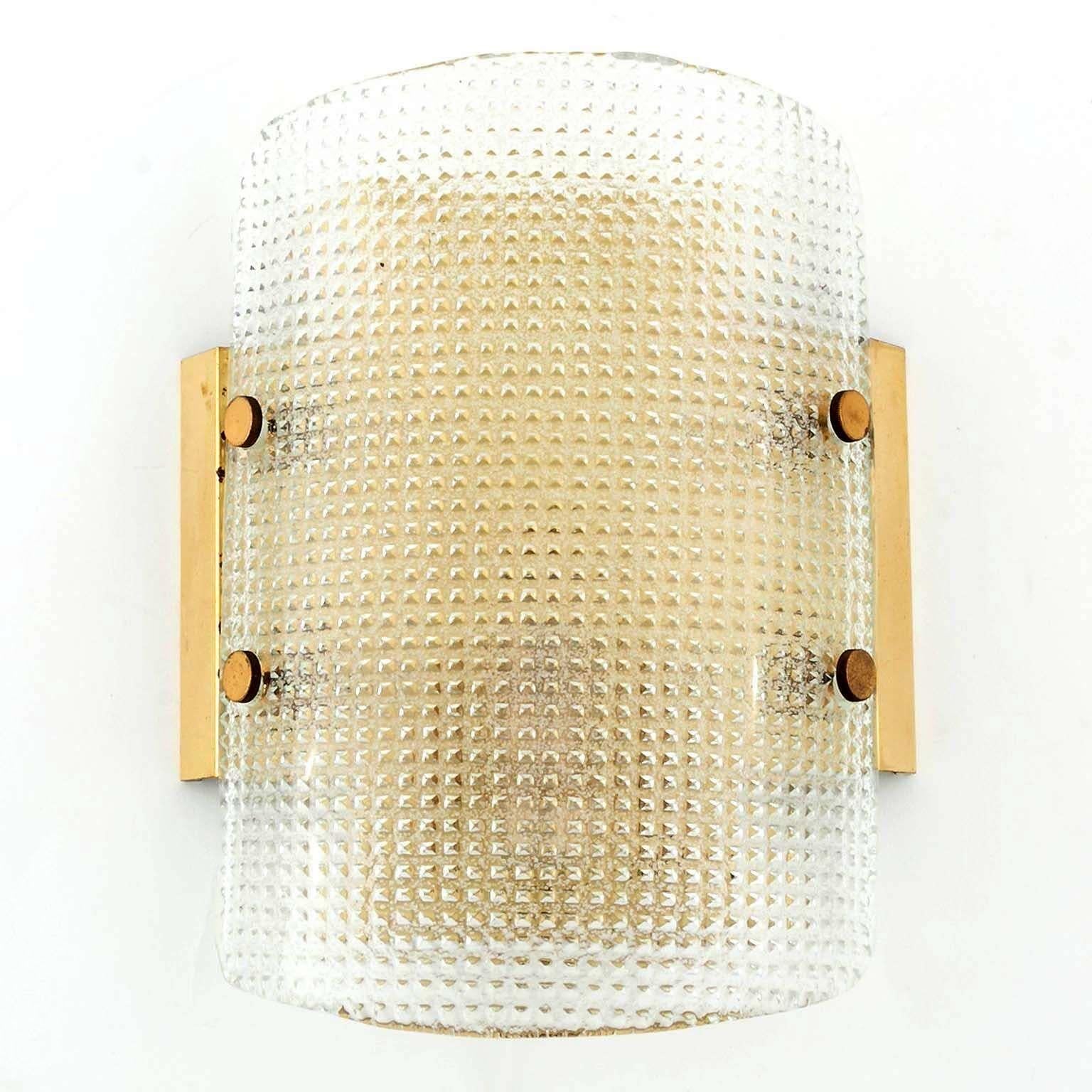A pair of wonderful wall lamps by Hillebrand, Germany, manufactured in midcentury, circa 1970 (late 1960s or early 1970s). 
A textured glass with a grid pattern is mounted with brass bolts on a polished brass backplate. Each fixture has one socket
