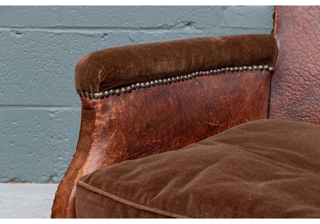 An older, likely European pair of leather club chairs with brass nail head details in an infinity motif on the wide crest rails. Large and smaller nail heads overall on the frames. The arms, seats and cushions upholstered in a brown velour like