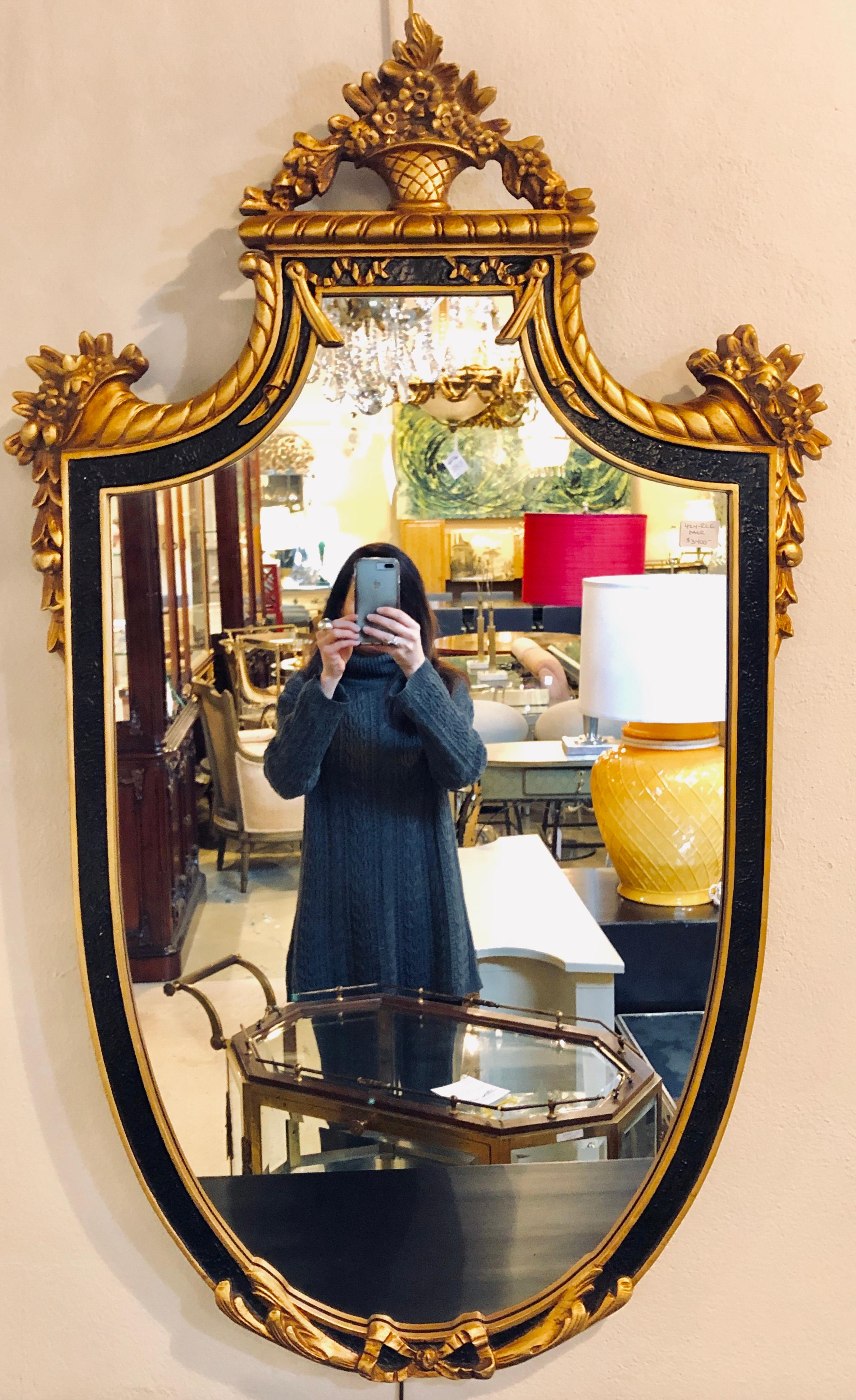 Pair of Hollywood Regency style ebony and giltwood wall or console mirrors. Simply spectacular are these Adams style wall or console mirrors. Each having an oval frame with a basket of flowers flowing onto a cornucopia all gilt on an ebonized and