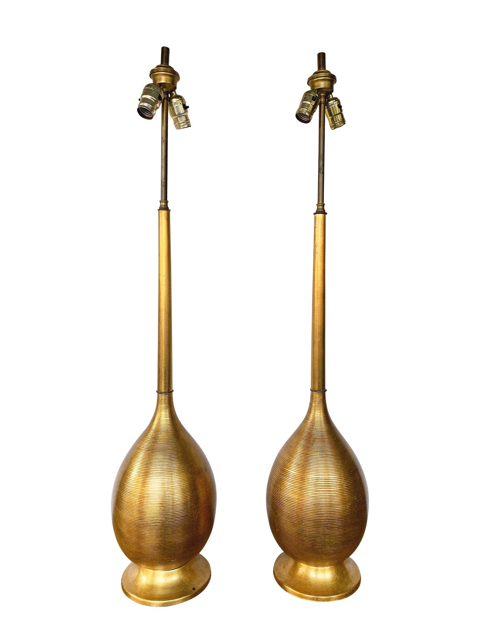 A pair of Hollywood Regency style lamps. They are comprised of a giltwood stem, gold-painted glass body, and gold-painted metal base with green felt lining. The lamps have double cluster bulb sockets and come with cream-white linen shades.