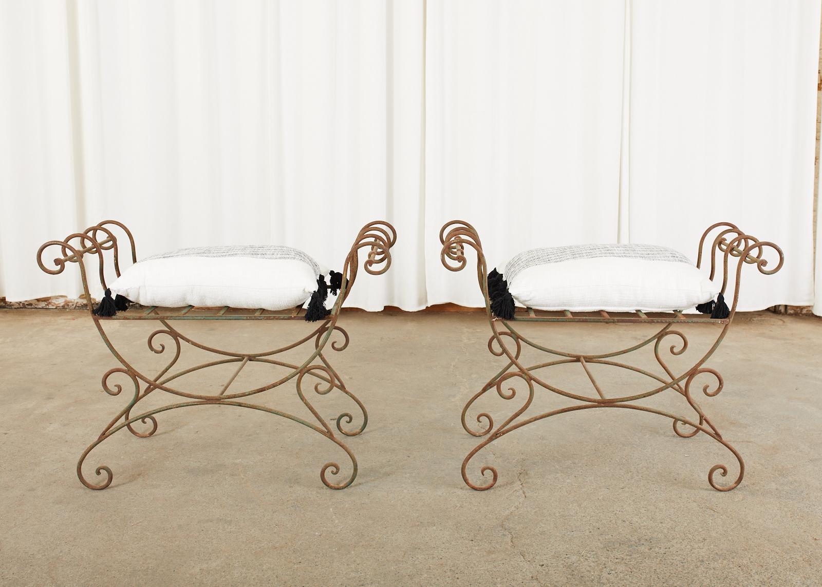 Stylish pair of Hollywood Regency wrought iron curule benches made in the neoclassical taste. Featuring an X-form design with scrolled arms and feet. The seats measure 25 inches wide and 18 inches deep. The wrought iron has a rich patinated metal