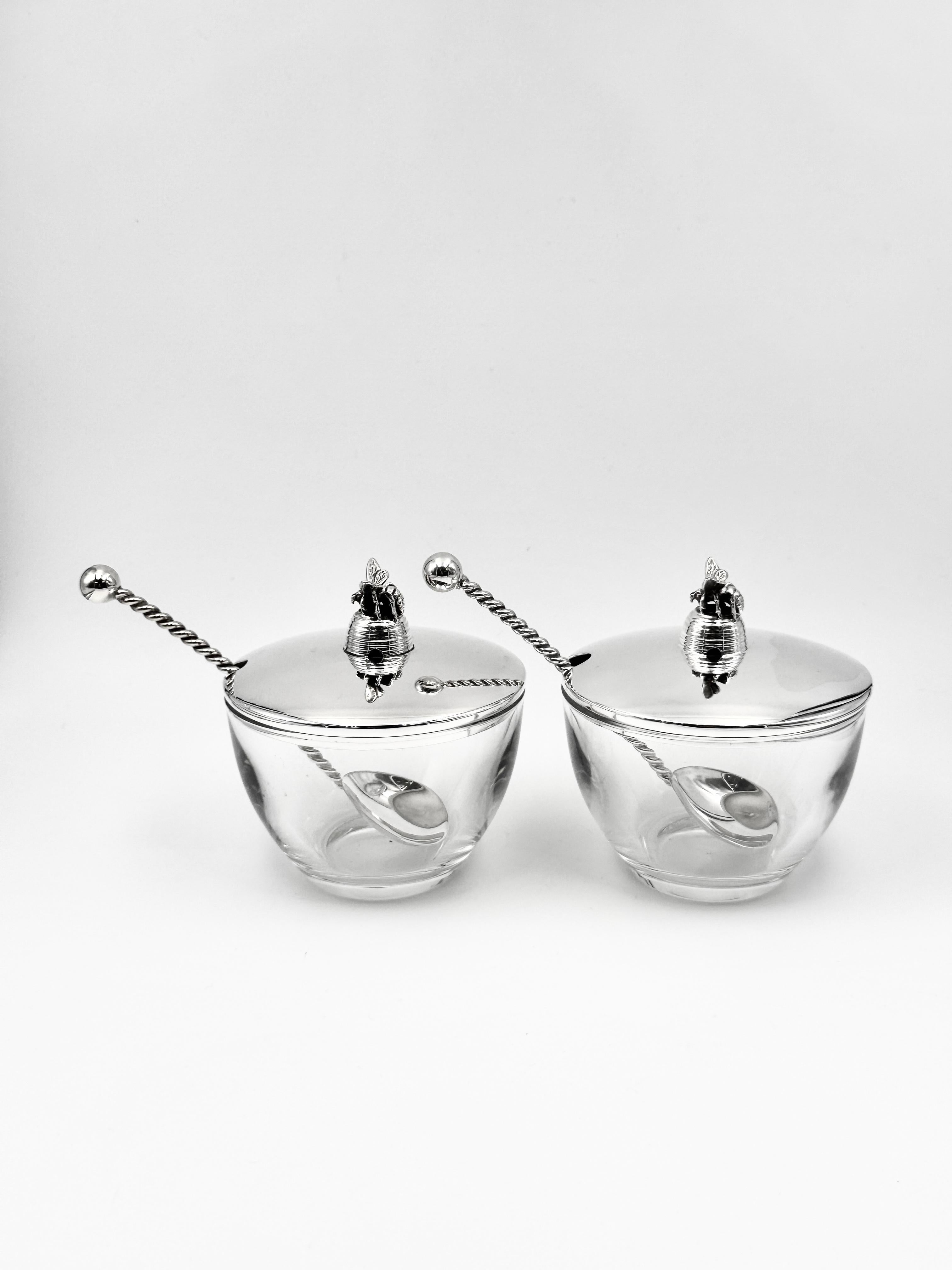 Two English sterling silver and glass honey pots with matching spoons, each fitted with a silver cover surmounted by a bee finial, hallmarked London 2003 and 2006 by Braybrook & Britten.
