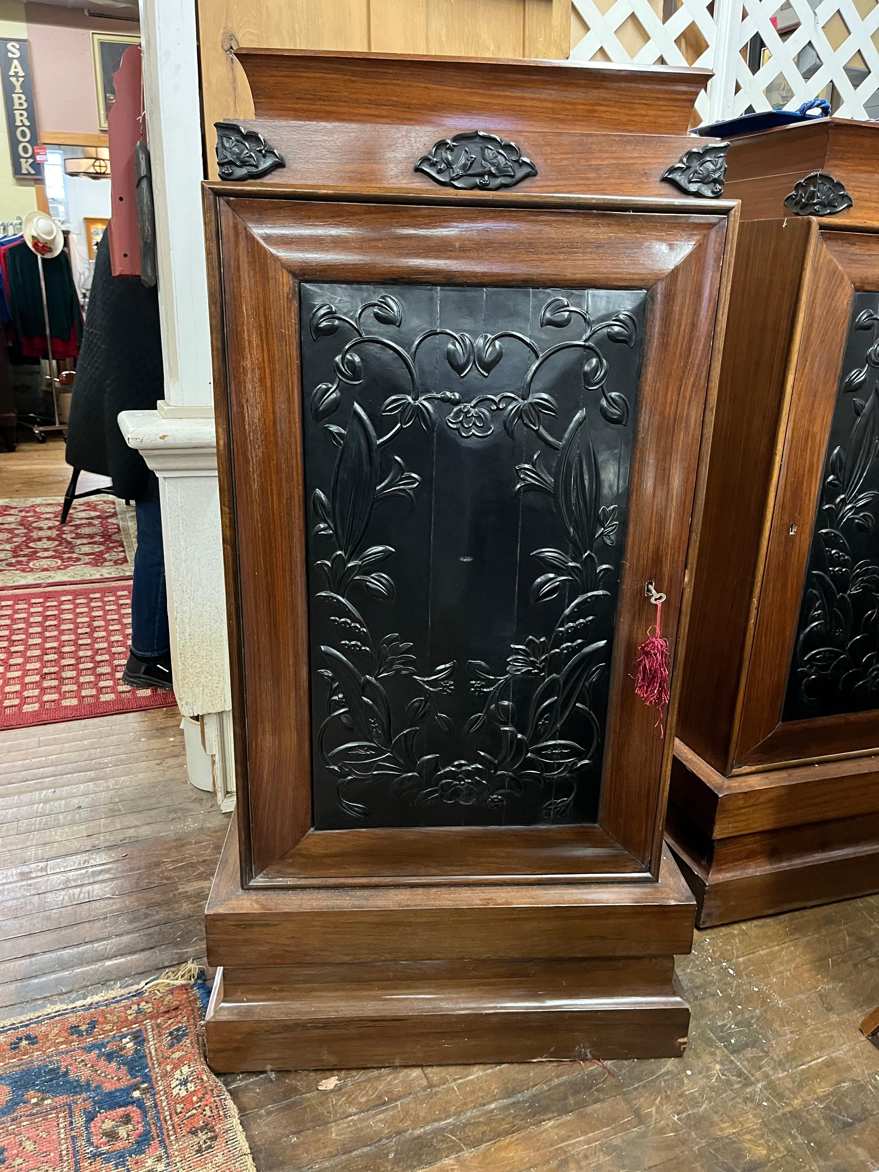 A decorative pair of Art Deco tall cabinets, circa 1930, in walnut with Asian inspired ebonized carved doors and decorative details, each on a plinth base. The interior of each retains its original adjustable shelf. Working locks and key. 