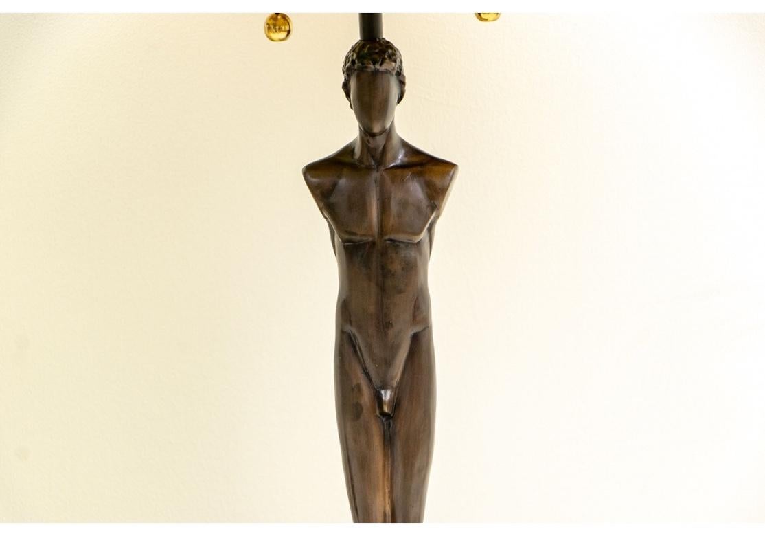 Designed by Tom Corbin (20th century sculptor). Patinated lost-wax bronze male and female standing nude figures as lamp supports, mounted on black marble bases. Signed on the bronze tiered bases. Flame form bronze finials. With black and gilt paper