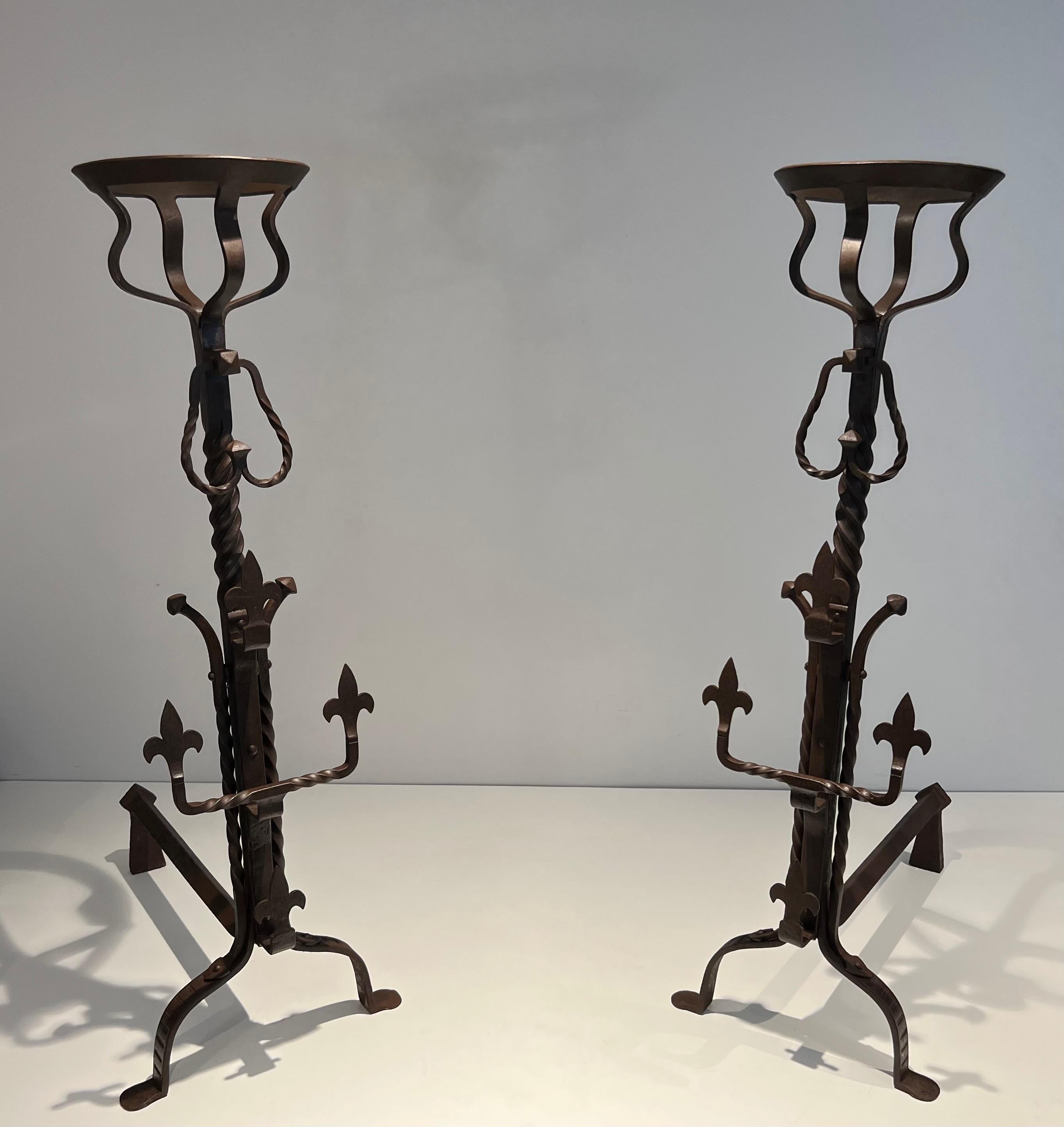 This tall pair of gothic style wrought iron andirons are also called landiers because of the shape of the top parts that where used to have water or soup boiling on receipt near the fire. They are rusty colored, have Fleurs de Lys decorations,