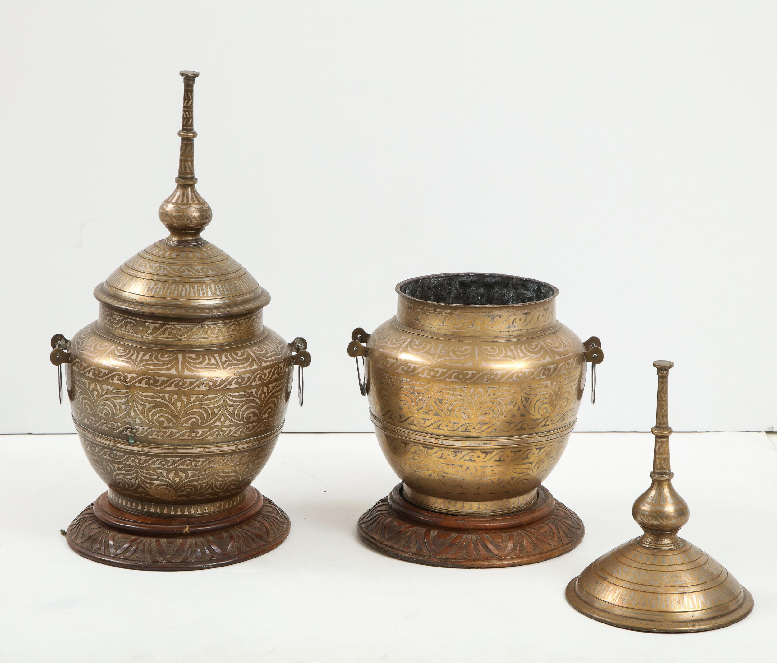 Attractive pair of mixed metal lidded jars, probably Indo-Persian 19th century, on carved wooden stands, having tall spire finials over turned bodies having pewter and other metals inlaid in an arabesque pattern.