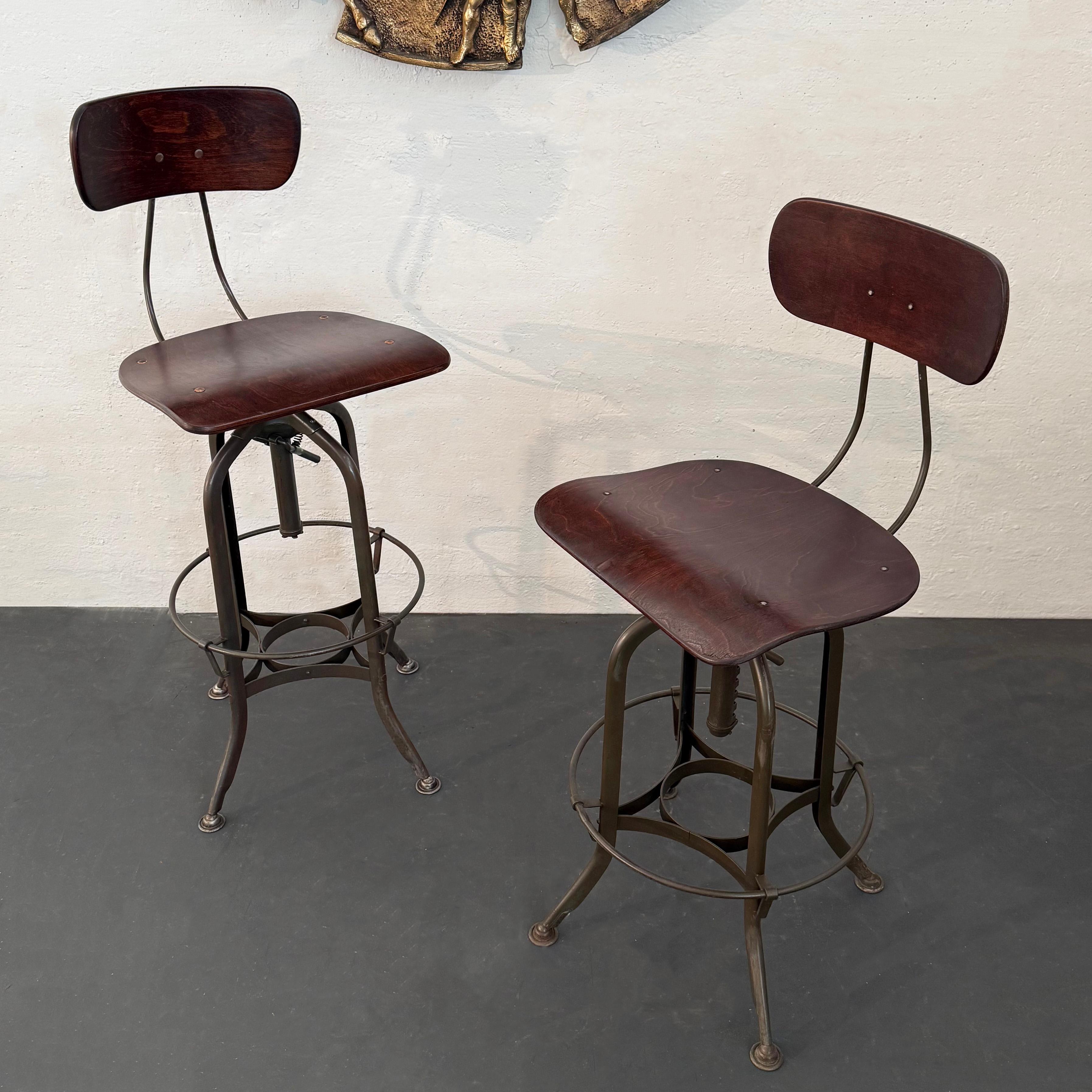 Pair of iconic, industrial drafting stools by Toledo Metal Furniture Co. circa 1940's feature height adjustable, painted steel star bases with bent maple ply, swivel seats and backs. The seat heights are adjustable from 28 - 33 inches. The footrests