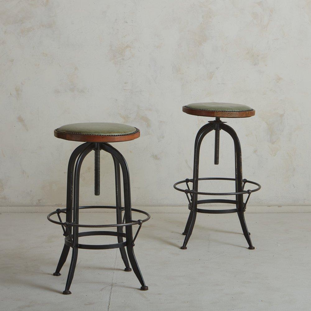 A pair of French 1950s industrial bar stools featuring adjustable, curved iron bases with footrests. These stools have circular wood seats in original green leather with stud detailing. Sourced in France, 1950s. The height of these stools is