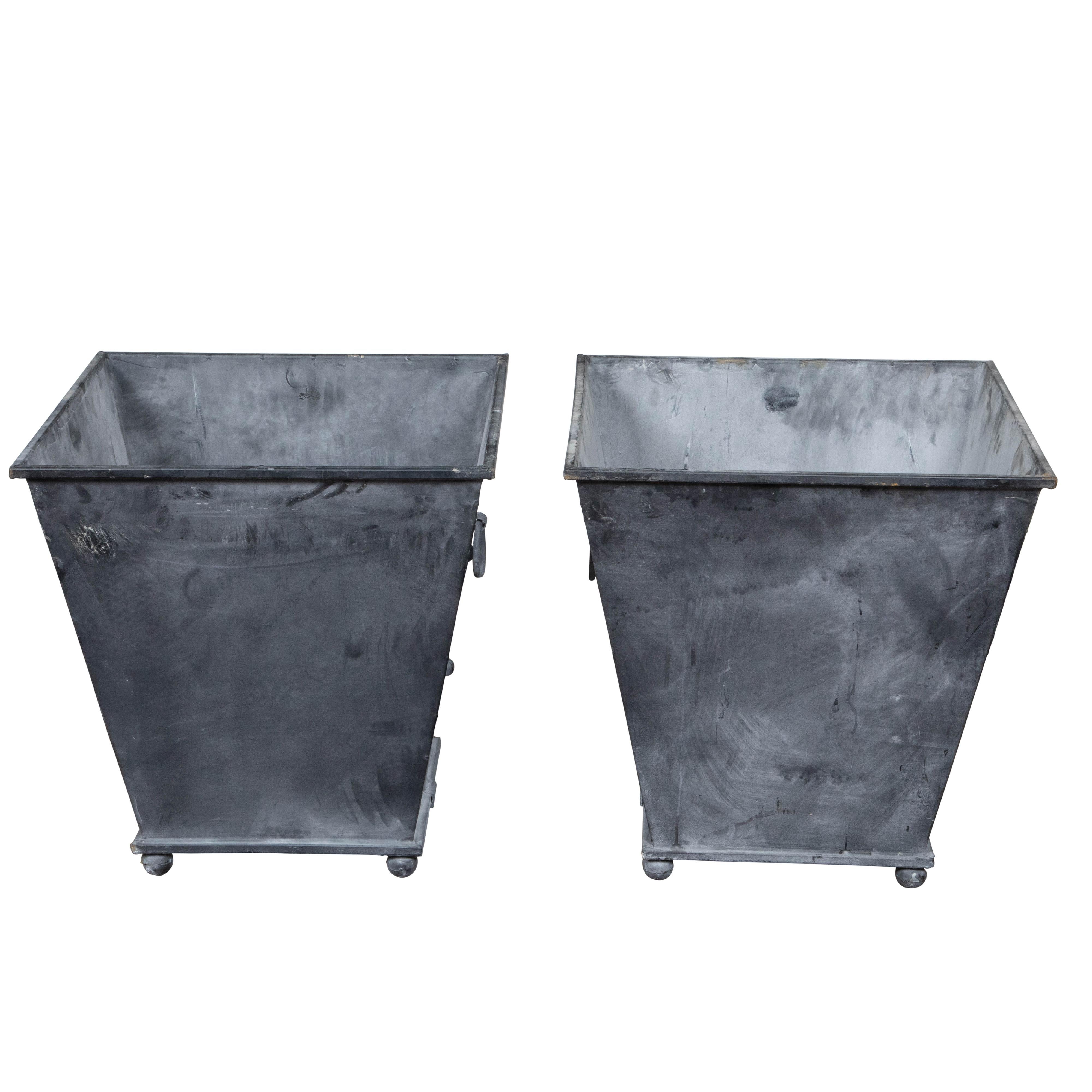 A pair of contemporary American metal cache pot planters from the 21st century with tapered lines, grey patinated color, lateral ring pulls and petite ball feet. Made in the USA during the 21st century, each of this pair of metal planters attracts