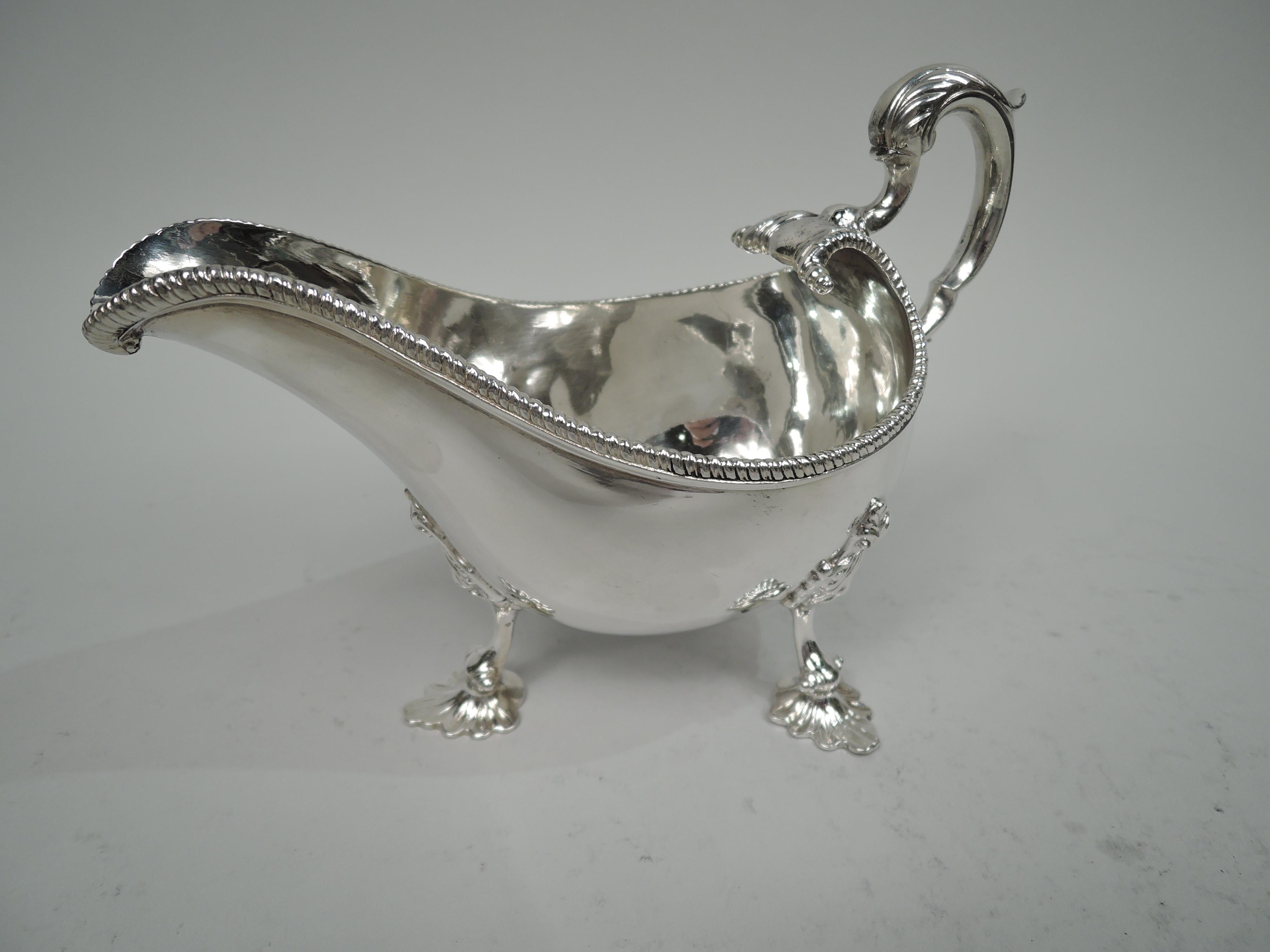 Pair of George III sterling silver gravy boats. Made by William Bond in Dublin in 1794. Each: Ovoid with helmet mouth and gadrooned rim. Leaf-capped double-scroll handle mounted to scrolled end. Three scallop-shell mounted leaf supports. Engraved
