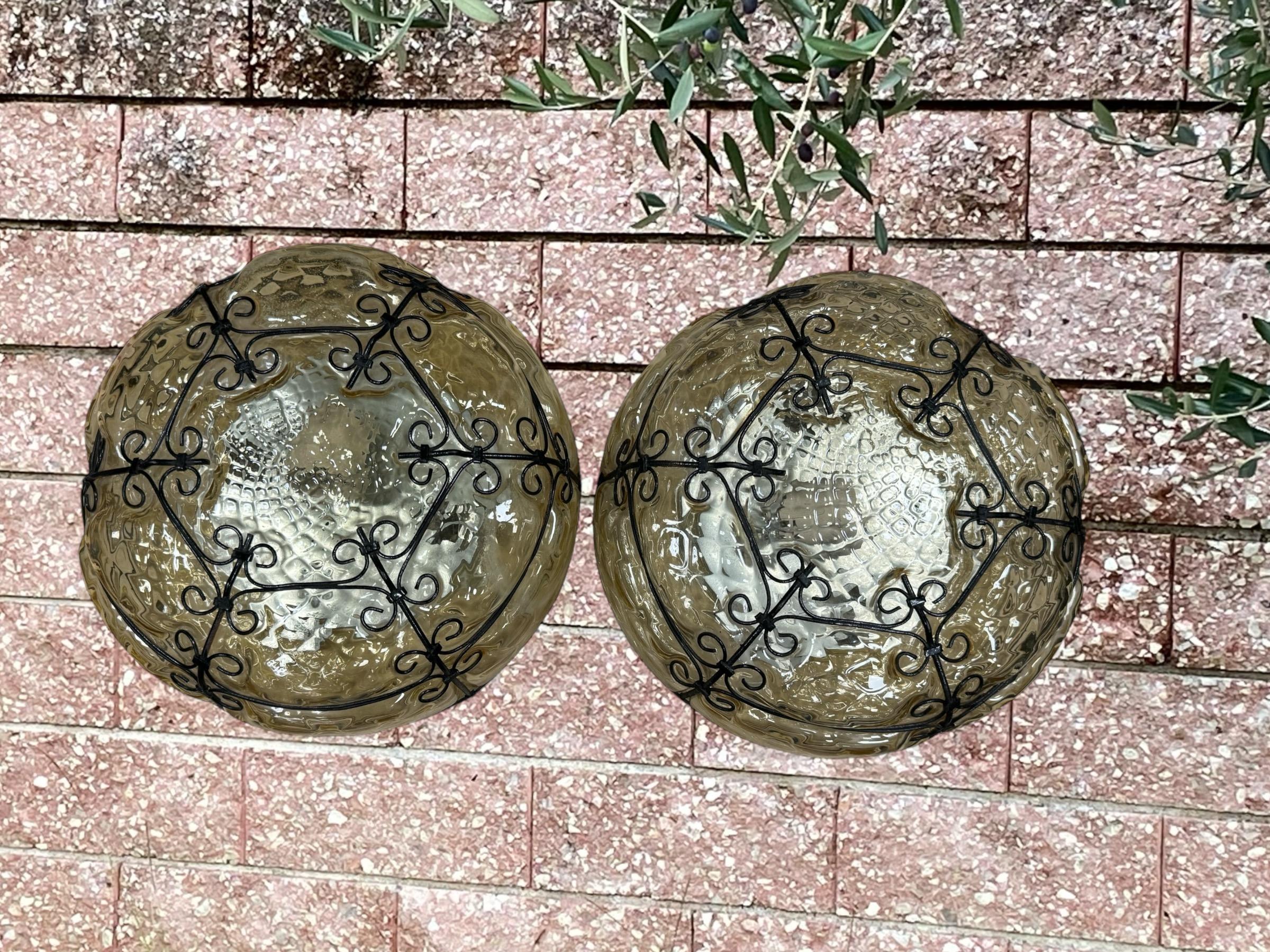 Un set di due vetri veneziani soffiati a mano e montati a filo, in vetro fumé o ambra, in una cornice di ferro. Può essere utilizzata anche come lampada da parete, in quanto il globo è fissato all'apparecchio con delle viti. È in condizioni vintage
