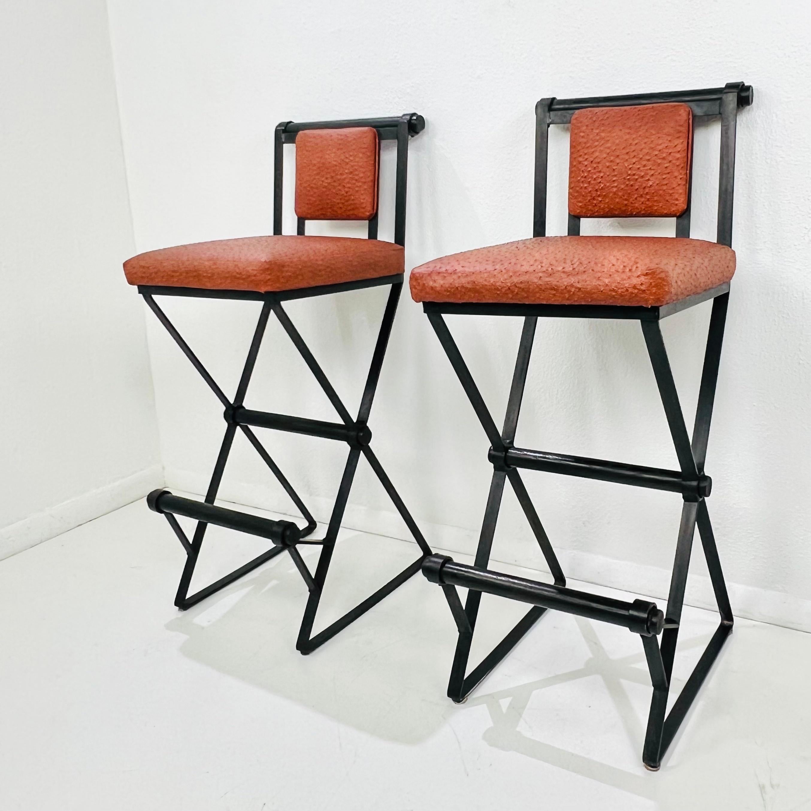 Une paire de tabourets de bar vintage, beaux et robustes, dans le style de Cleo Baldon. Structure en fer forgé avec revêtement en poudre noir, assise et dossier en cuir d'autruche de couleur terre de sienne brûlée. Les barres arrondies en bois de