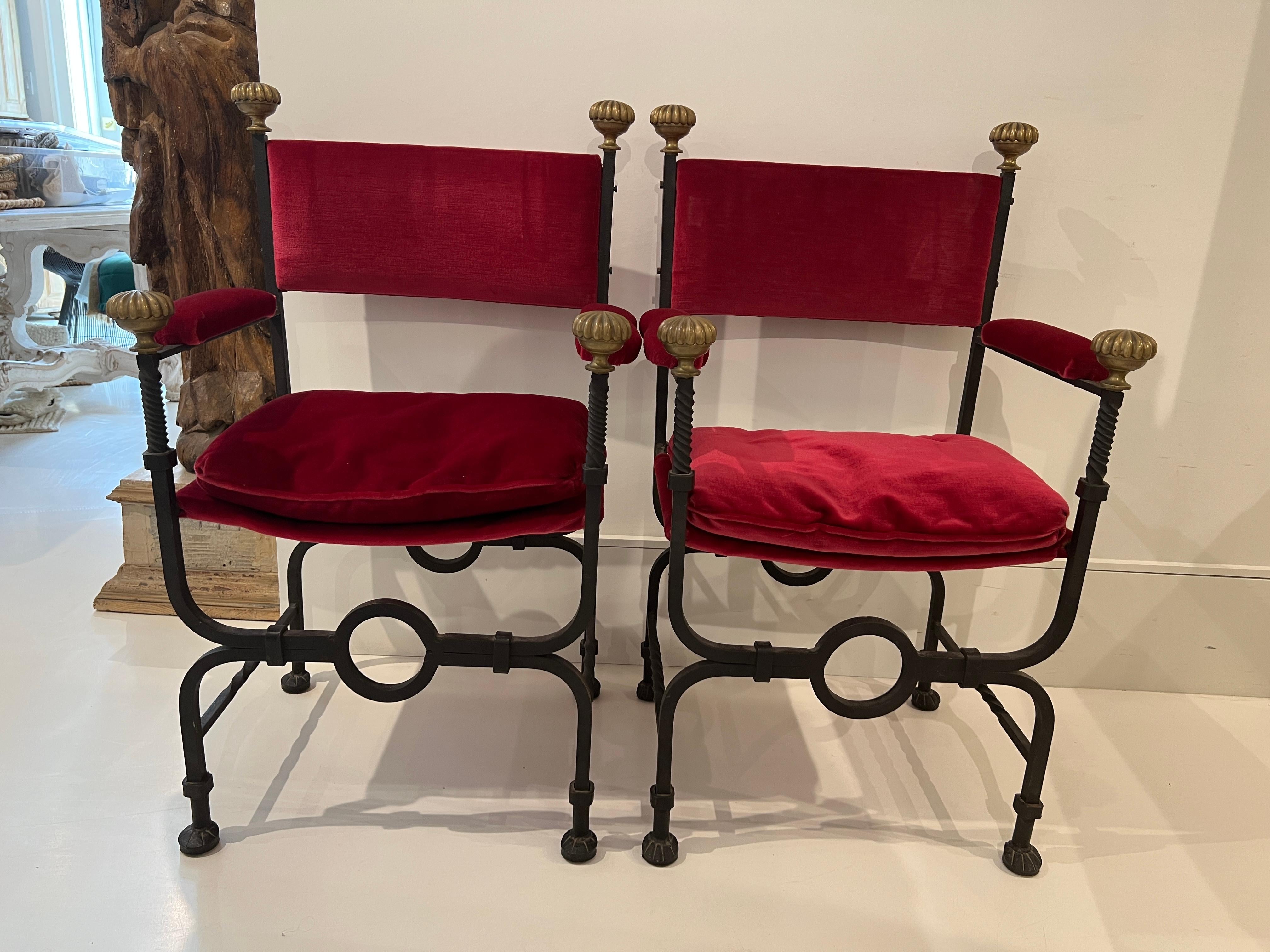 A pair of Savonarola chairs fabricated in heavy iron with brass embellishments. The seats, arms and back are upholstered in lush red velvet.
