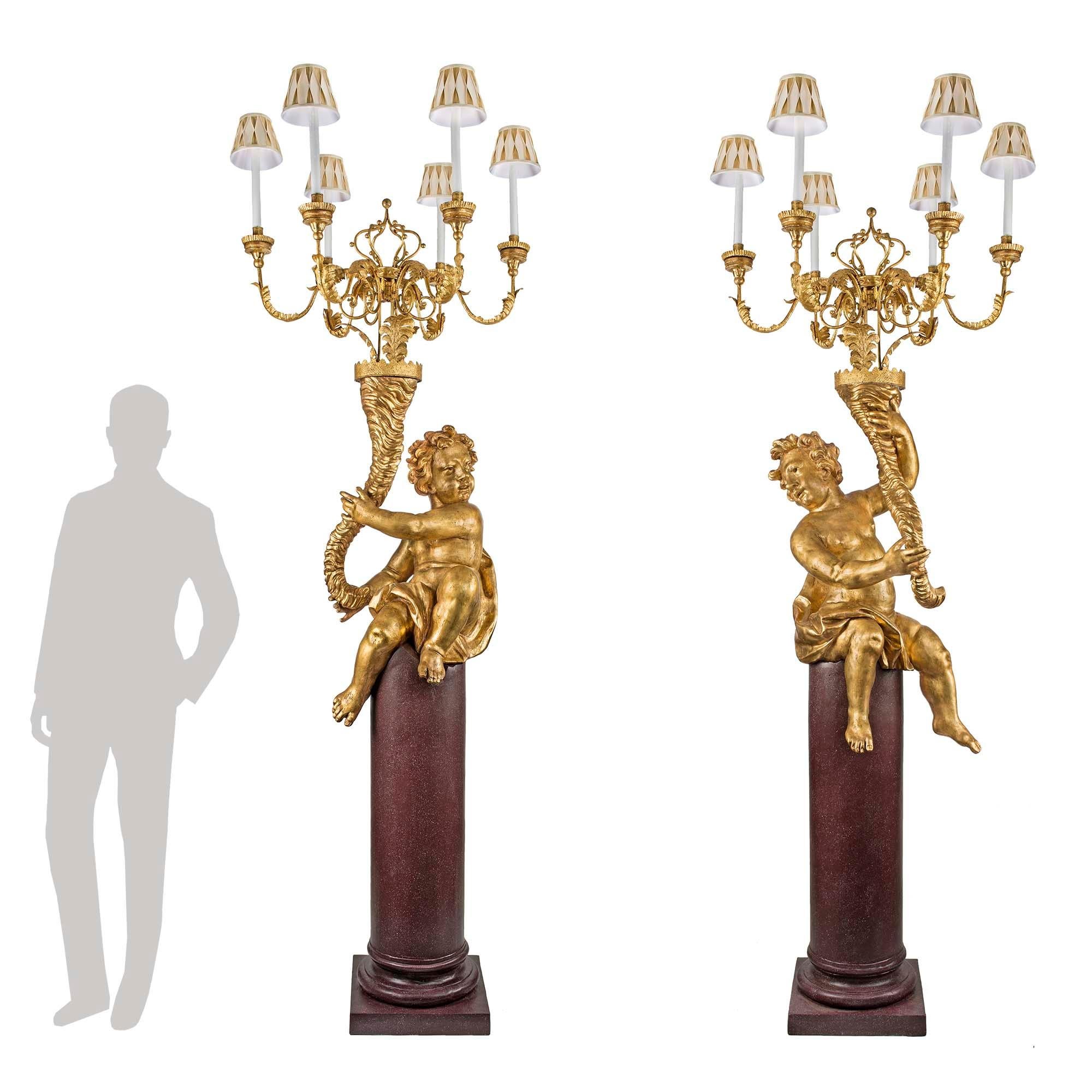 A stunning and monumental true pair of Italian 17th century giltwood and gilt iron six arm cherub candelabra torchières, circa 1680. Each torchières is raised by a sensationally 19th century faux painted Porphyry pedestal, with a mottled bottom