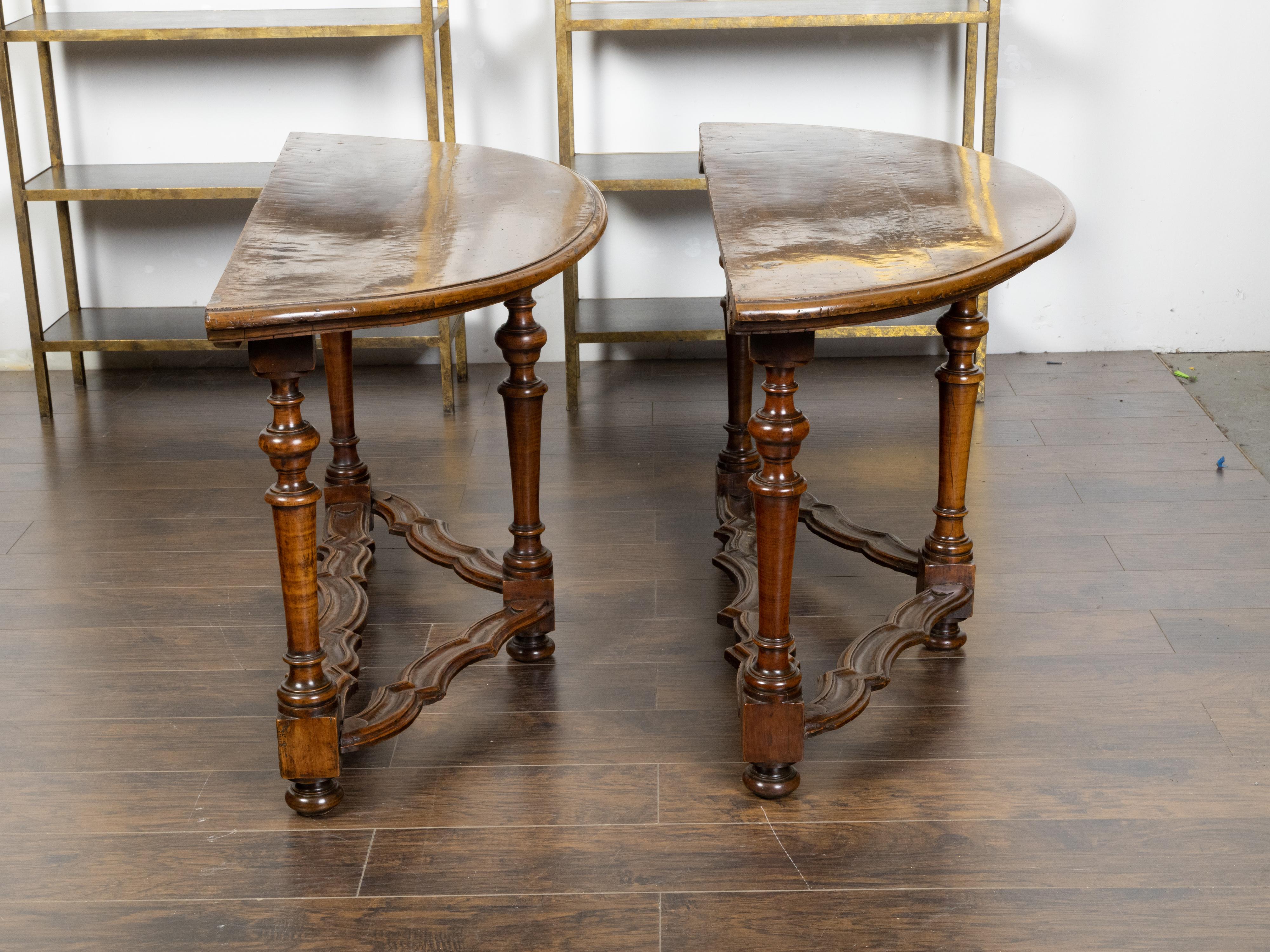 A pair of Italian Baroque style walnut demi-lune console tables from the early 19th century, with semi-circular tops, turned legs and carved stretchers. Created in Italy during the first quarter of the 19th century, each of this pair of demi-lune