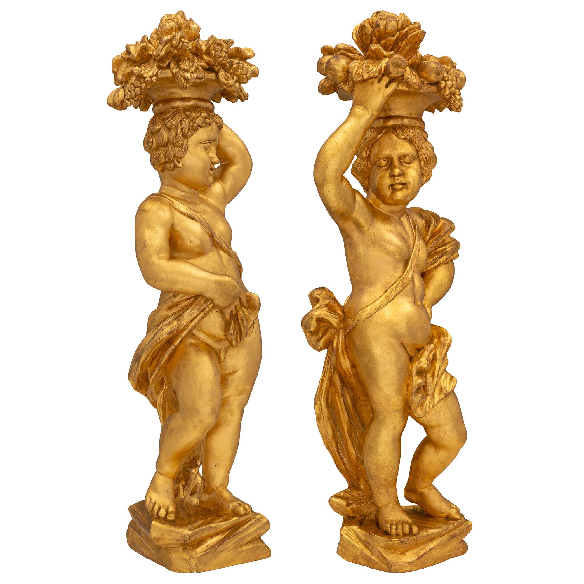 A stunning and extremely decorative true pair of Italian 18th century Baroque period giltwood statues. Each statue is raised by a rectangular ground like designed base where the charming cherubs stand. Each is draped in wonderfully executed flowing