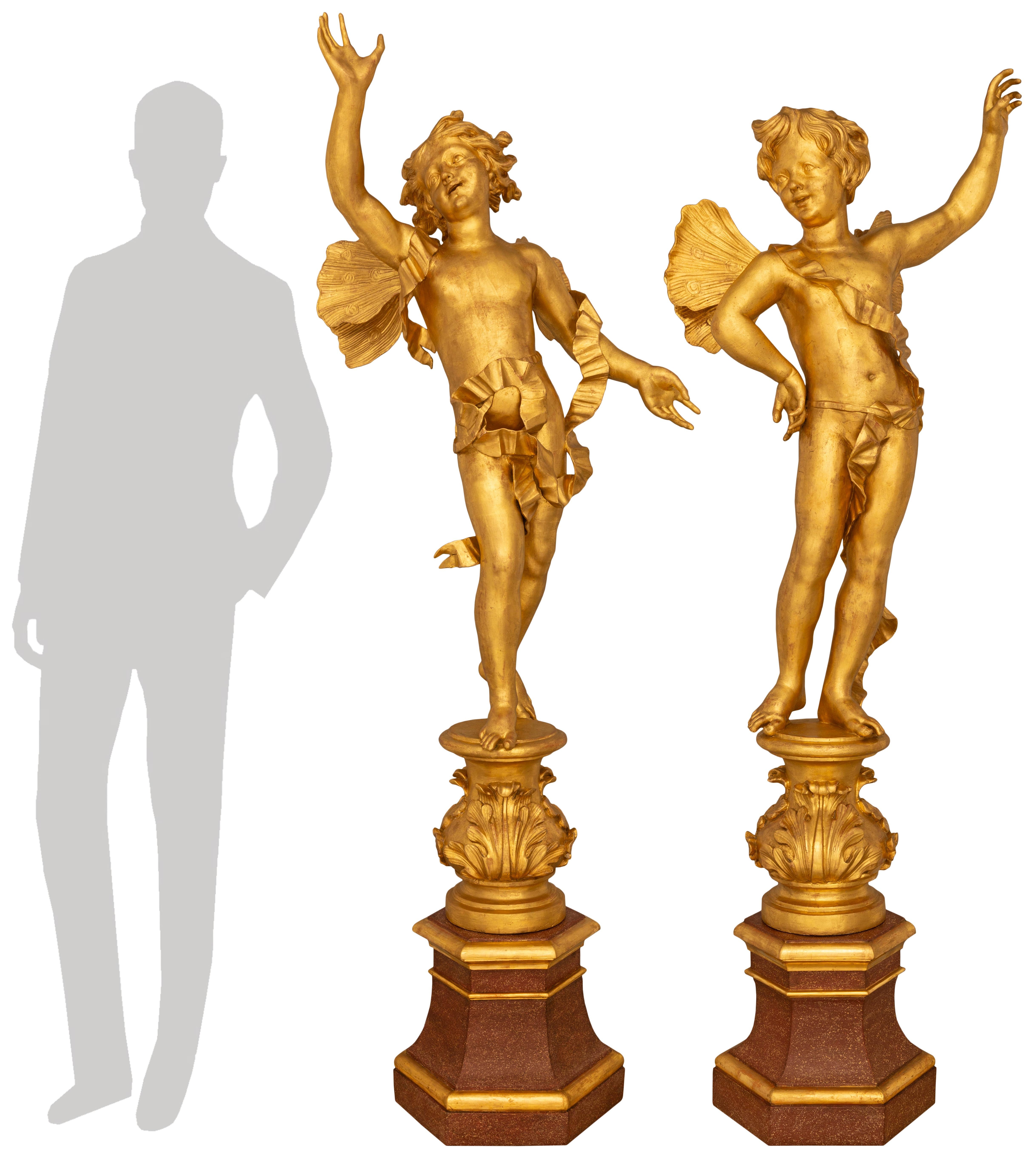 Un'impressionante e affascinante coppia di statue barocche italiane del XVIII secolo in finto marmo dipinto e legno dorato. Entrambe le allegre statue sono sollevate su piedistalli esagonali affusolati in finto marmo dipinto con bande di Giltwood