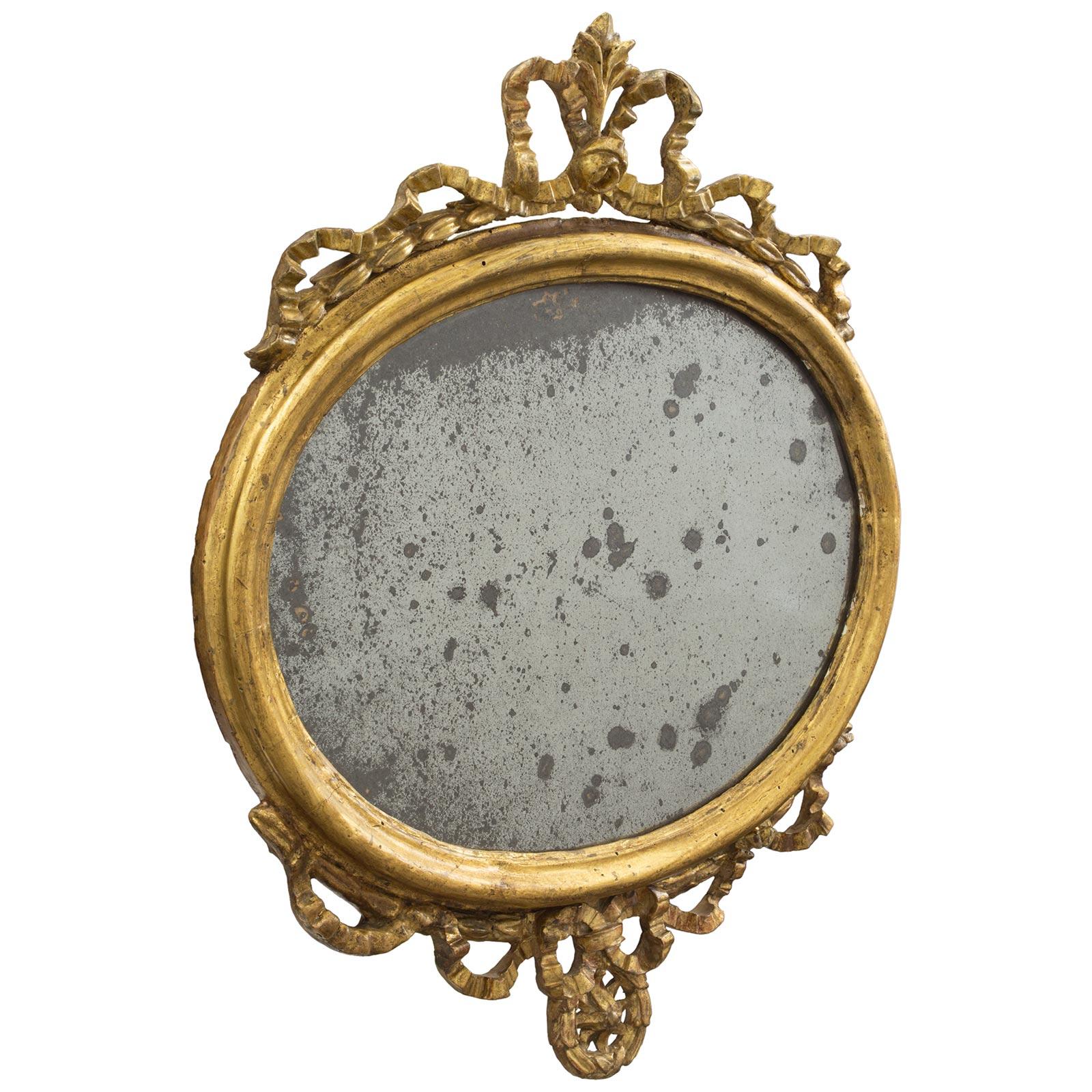 A charming pair of Italian 18th century Louis XVI period giltwood mirrors. Each mirror frame has a mottled oval frame with an elegant bottom reserve of a swaging ribbon with a central tied bow over a laurel wreath centered by a rosette. The top