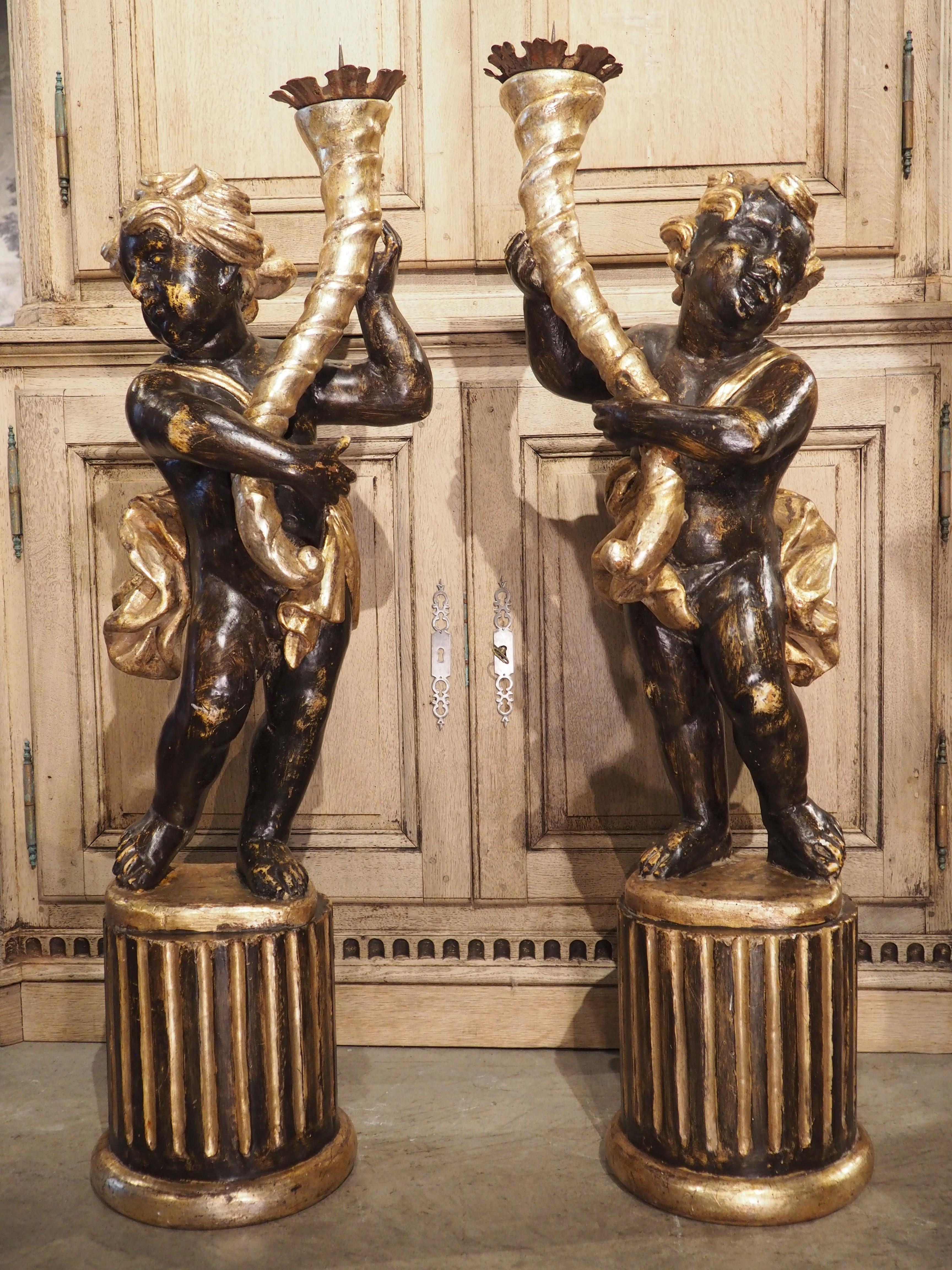 Standing at just under 4 feet high and hand-carved in Italy during the 1700’s, this fabulous pair of hand-painted and gilt putti torcheres have sinuous cornucopia columns. Each putto is depicted standing contrapposto on top of a brown Doric order