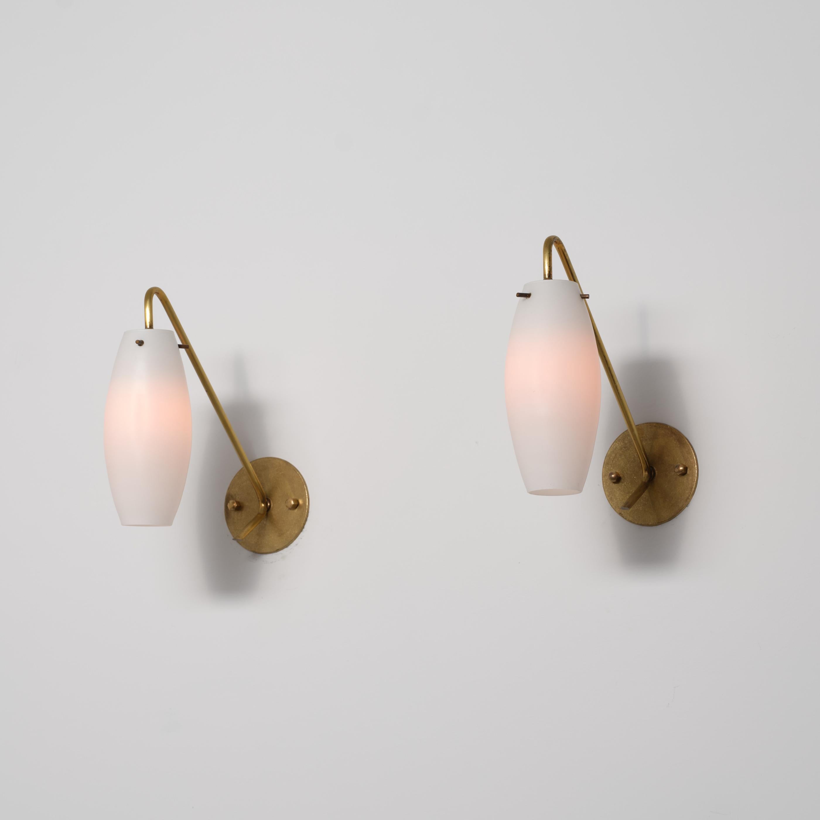 Pareja de apliques de pared italianos de los años 50, que combinan un diseño modernista minimalista con refinados detalles de latón. Cada lámpara presenta una pantalla alargada de cristal opalino con una silueta suave y cónica, suspendida de un