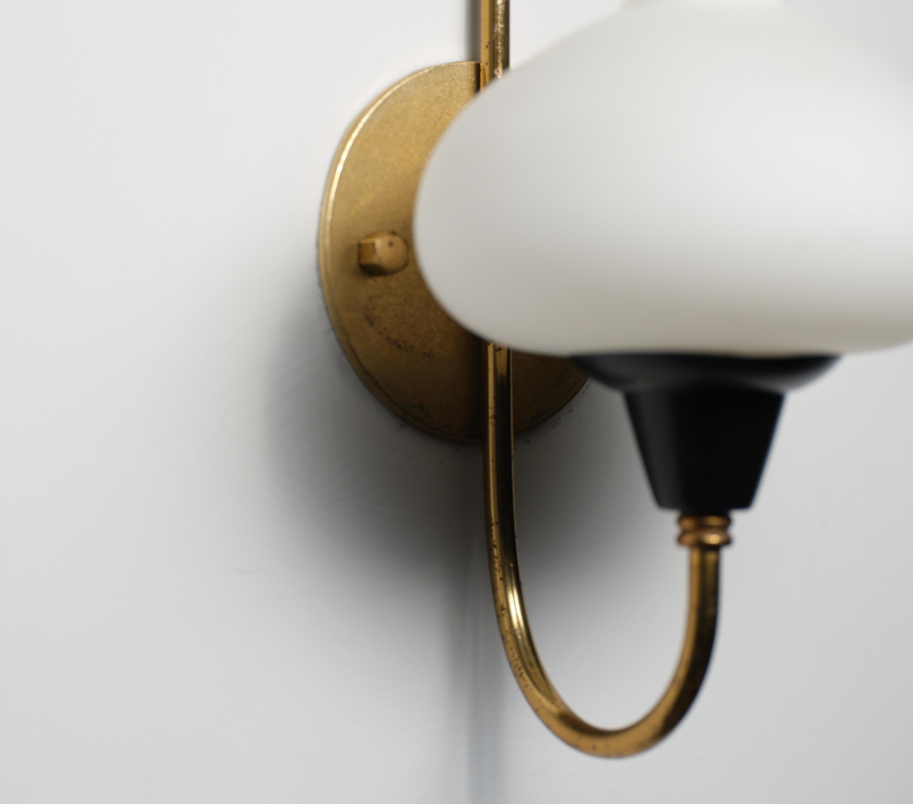 Coppia di appliques prodotte in Italia negli anni Cinquanta. Ogni applique è composta da una piastra posteriore in ottone orientata verticalmente, un braccio in ottone curvo con un supporto per la presa in metallo laccato nero e un diffusore in