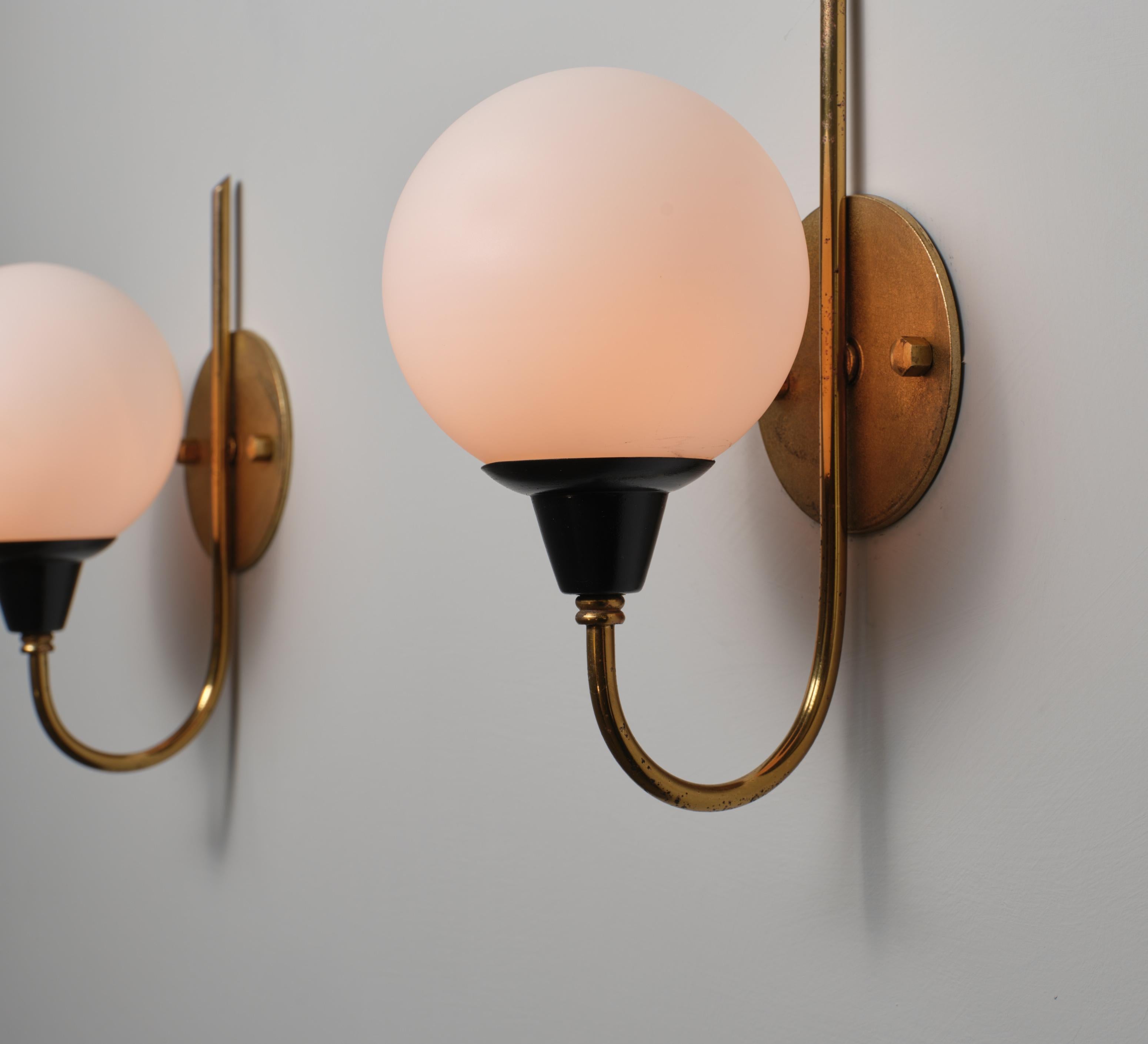 Pair of wall sconces designed and manufactured in Italy during the 1950s. The structure includes a vertical brass backplate with an integrated rod, a U-shaped brass arm, and a black lacquered metal socket mount. Each fixture supports a spherical