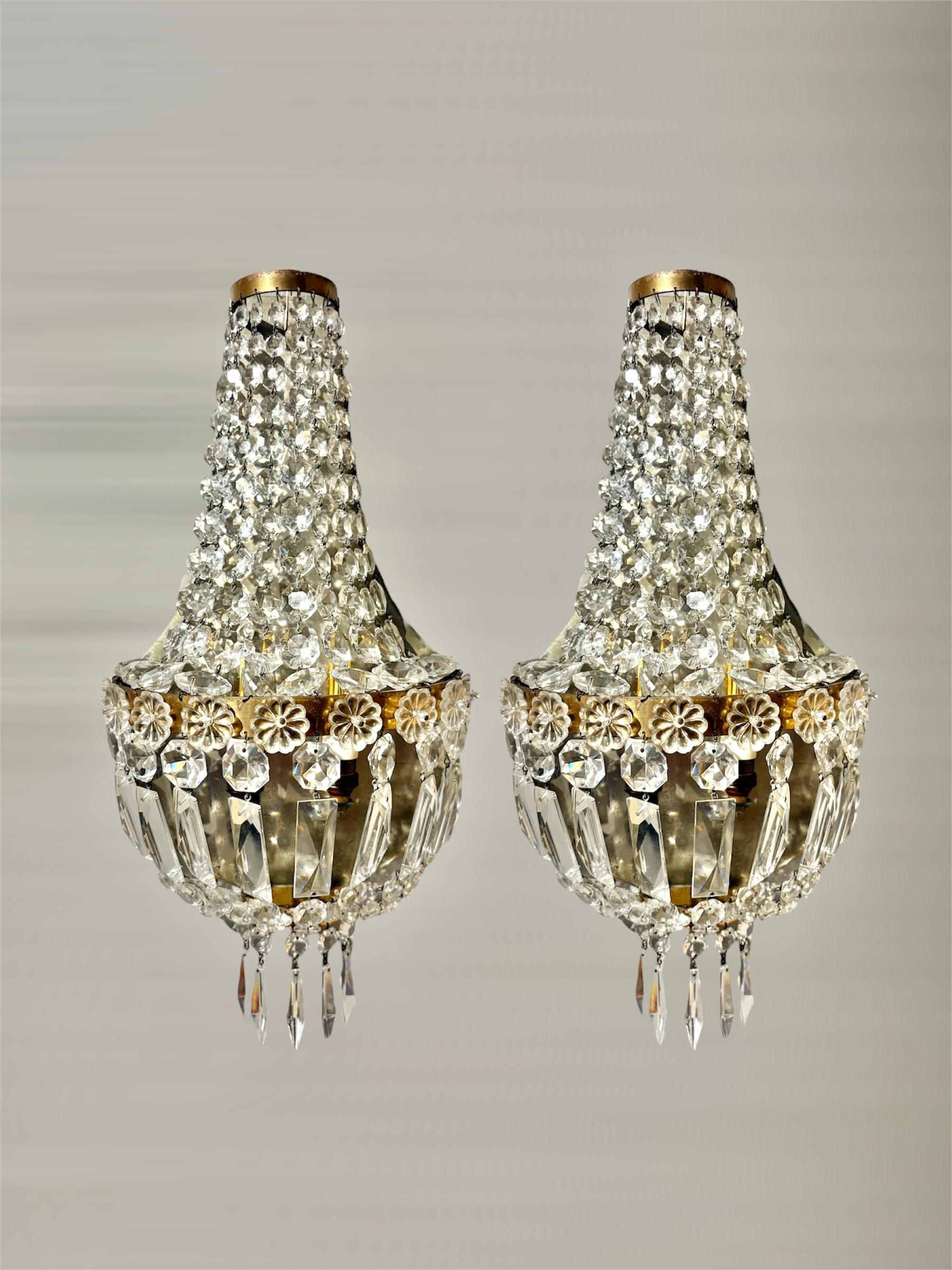 Questa elegante coppia di applique in stile Eleg risale agli anni Cinquanta. Ogni pezzo è riccamente ornato da prismi di cristallo a cascata che creano un effetto raffinato e luminoso. Il design è caratterizzato da una forma affusolata, che si