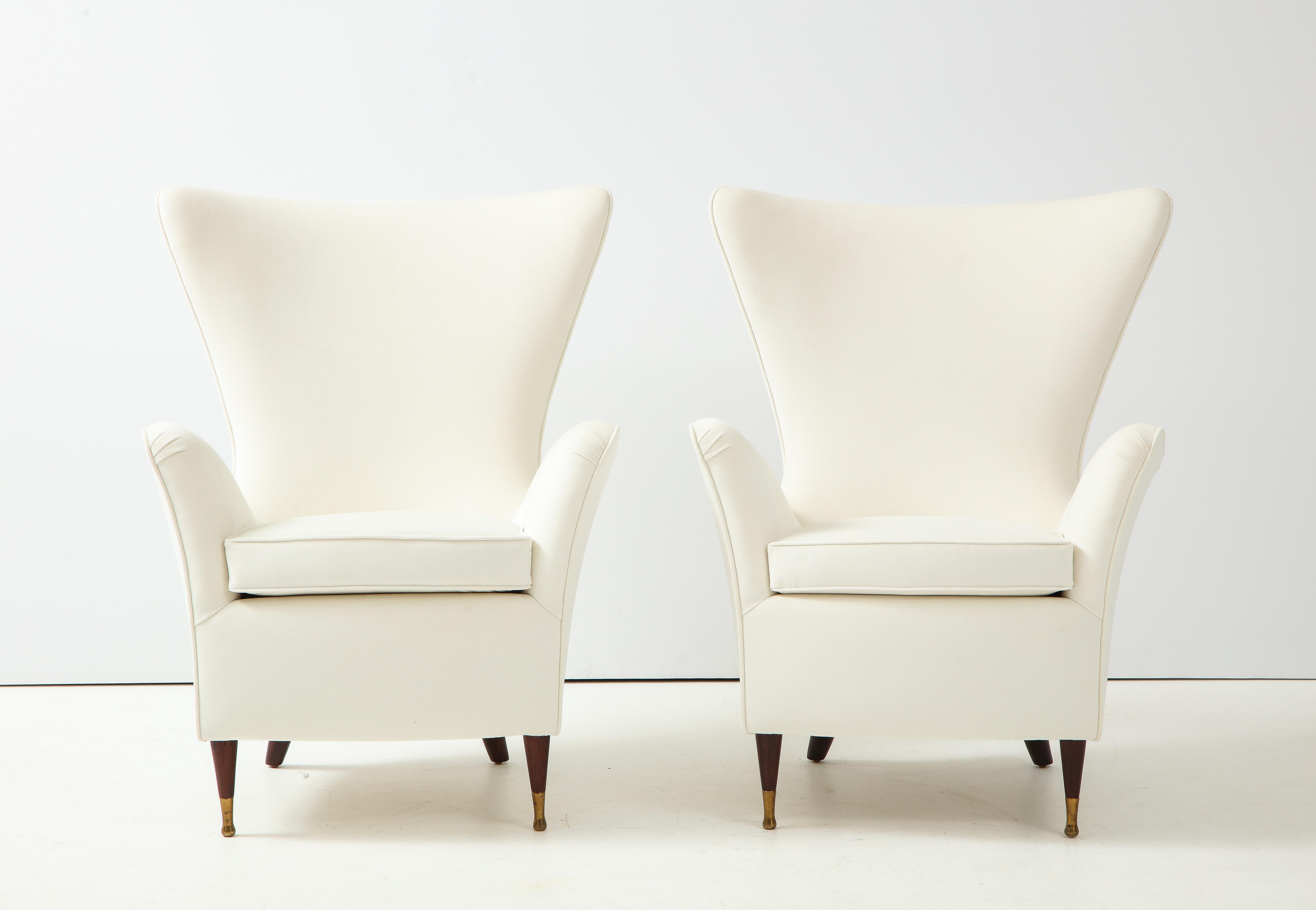 A pair of sculptural Italian 1950's wing high back arm chairs; supported on elegant walnut legs the front straight and ending in brass sabot and the back sabre shaped. The strong lines and proportions of the chairs are perfectly balanced, with the