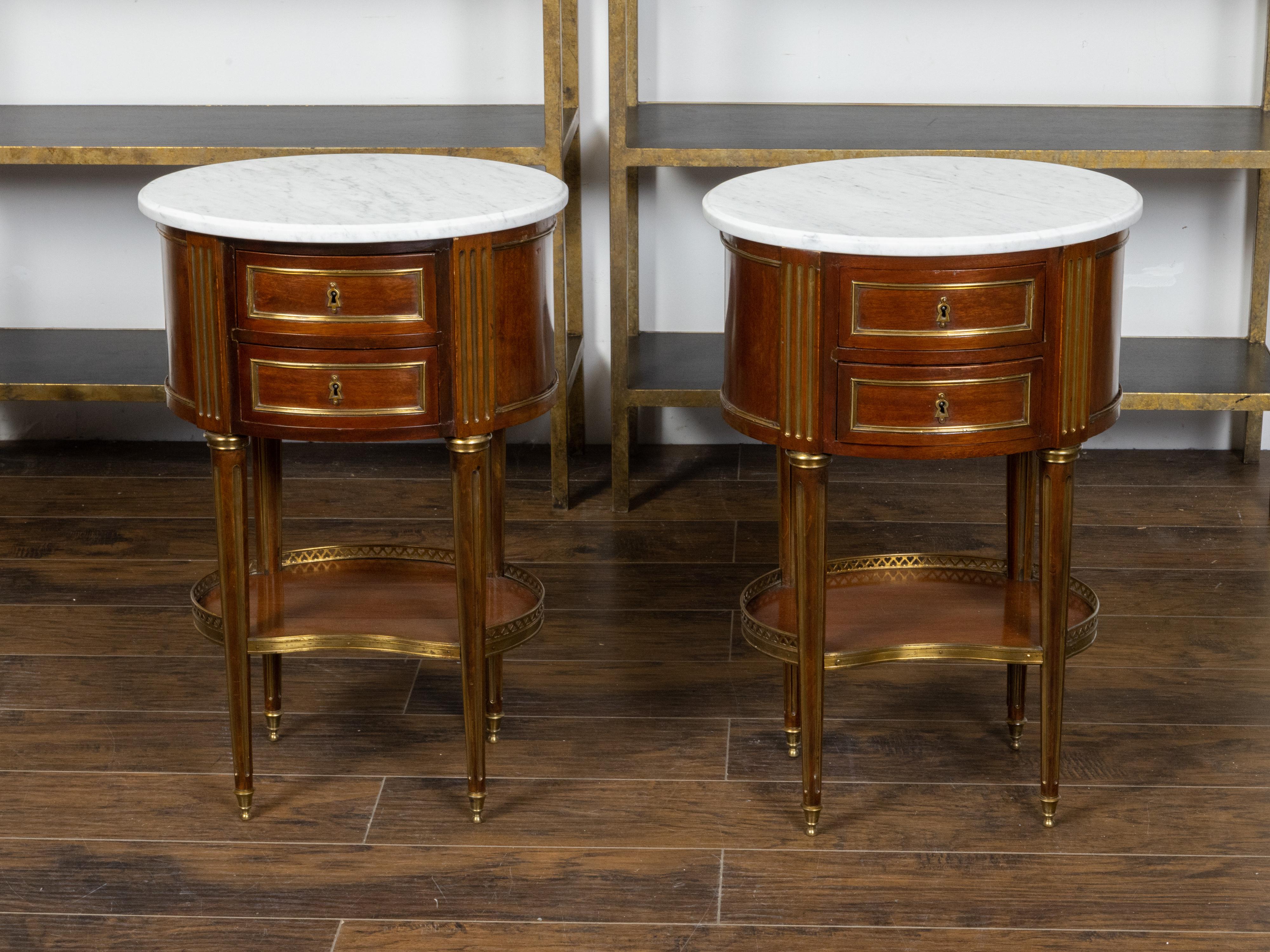 A pair of Italian neoclassical style mahogany bedside tables from the 19th century, with white oval marble tops, two drawers, gilt fluted accents, brass galleries and bean-shaped shelves. Created in Italy during the 19th century, each of this pair