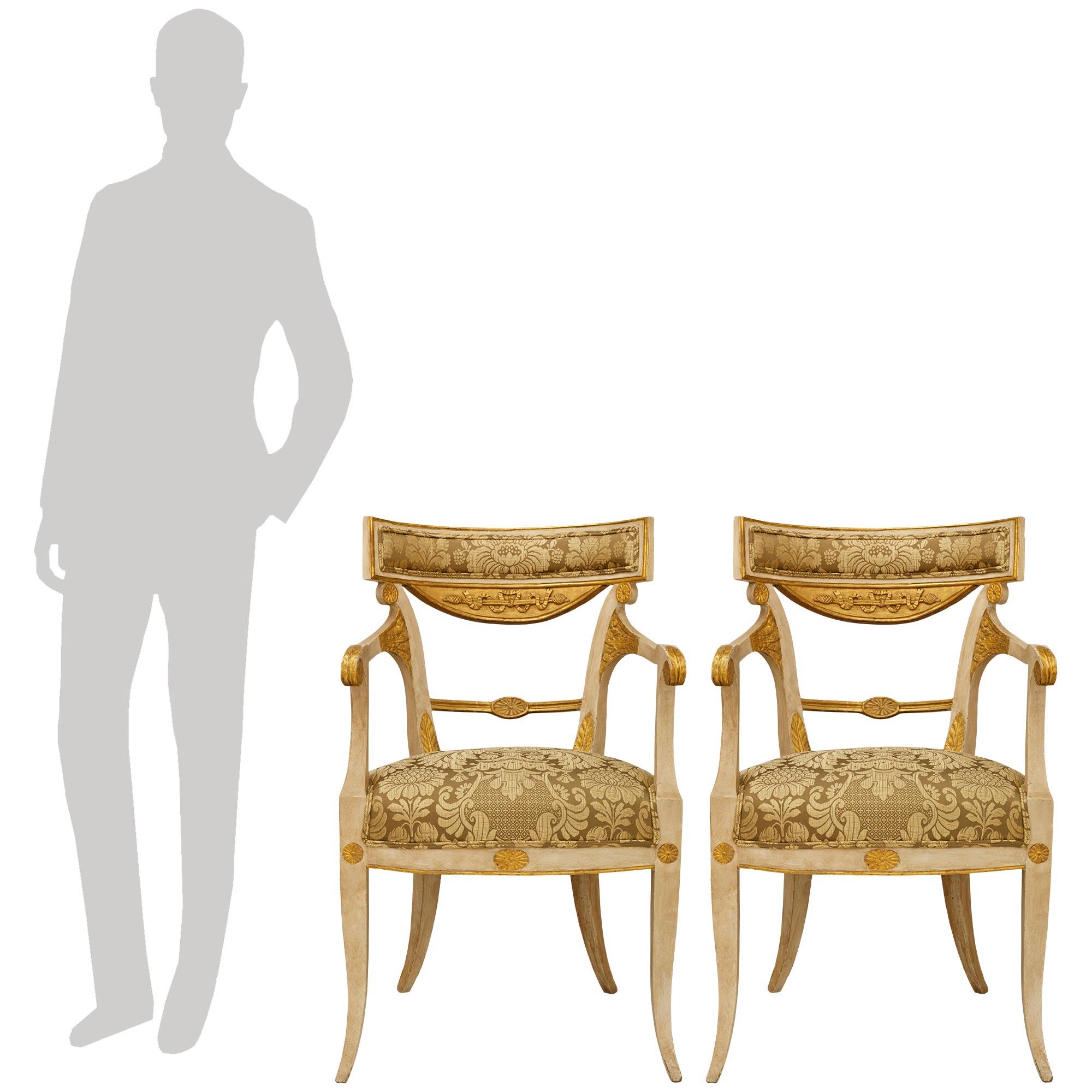 Une élégante et charmante paire de fauteuils néoclassiques italiens du début du 19e siècle en bois patiné et doré. Chaque belle chaise repose sur des pieds carrés courbés et effilés, rehaussés de rosettes en bois doré au-dessus de chaque pied et au