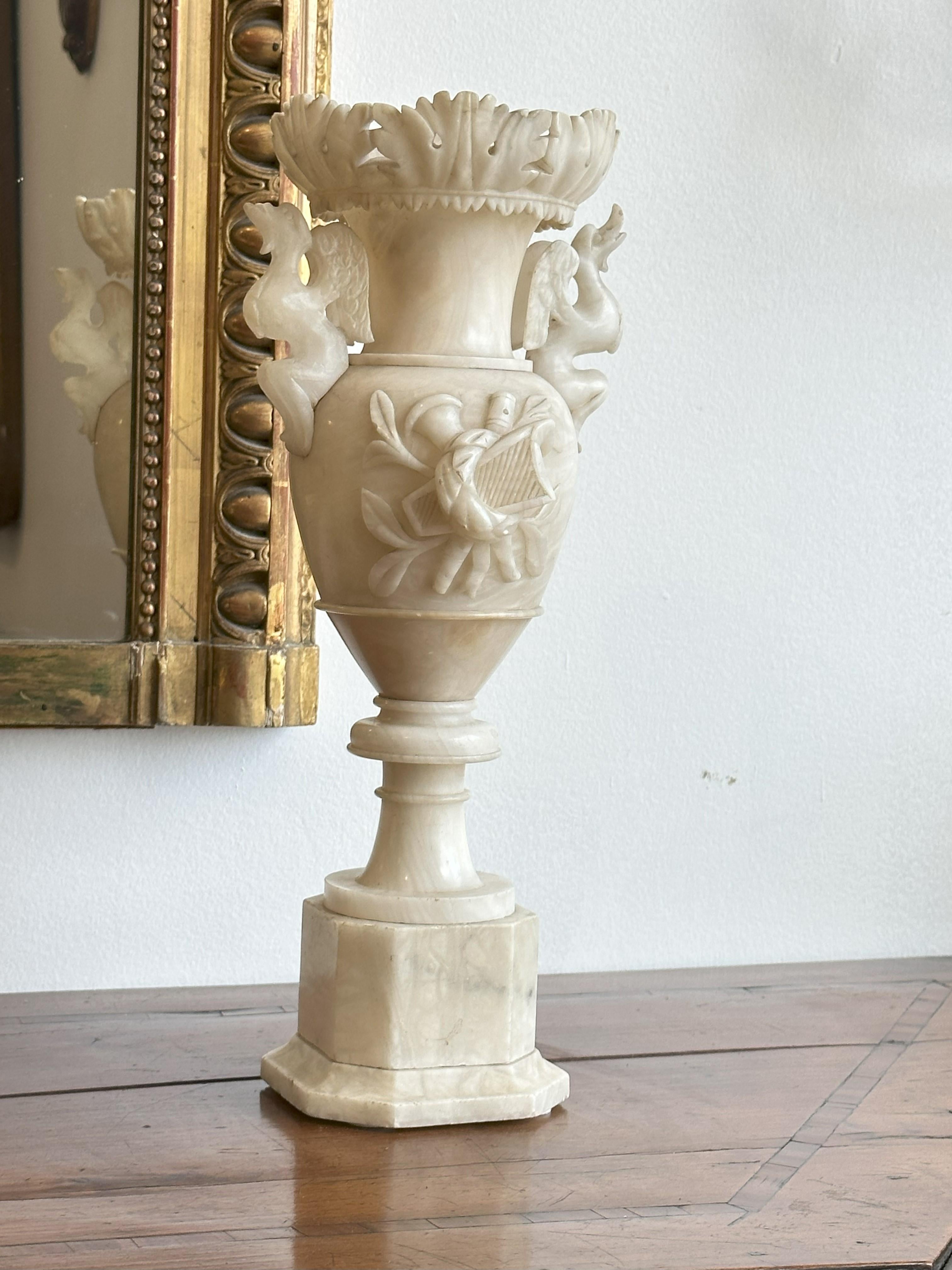 Each of baluster form urn surmounted by a final of scrolling foliage and flanked by carved griffin-form handles raised on a circular socle over an octagonal plinth base.