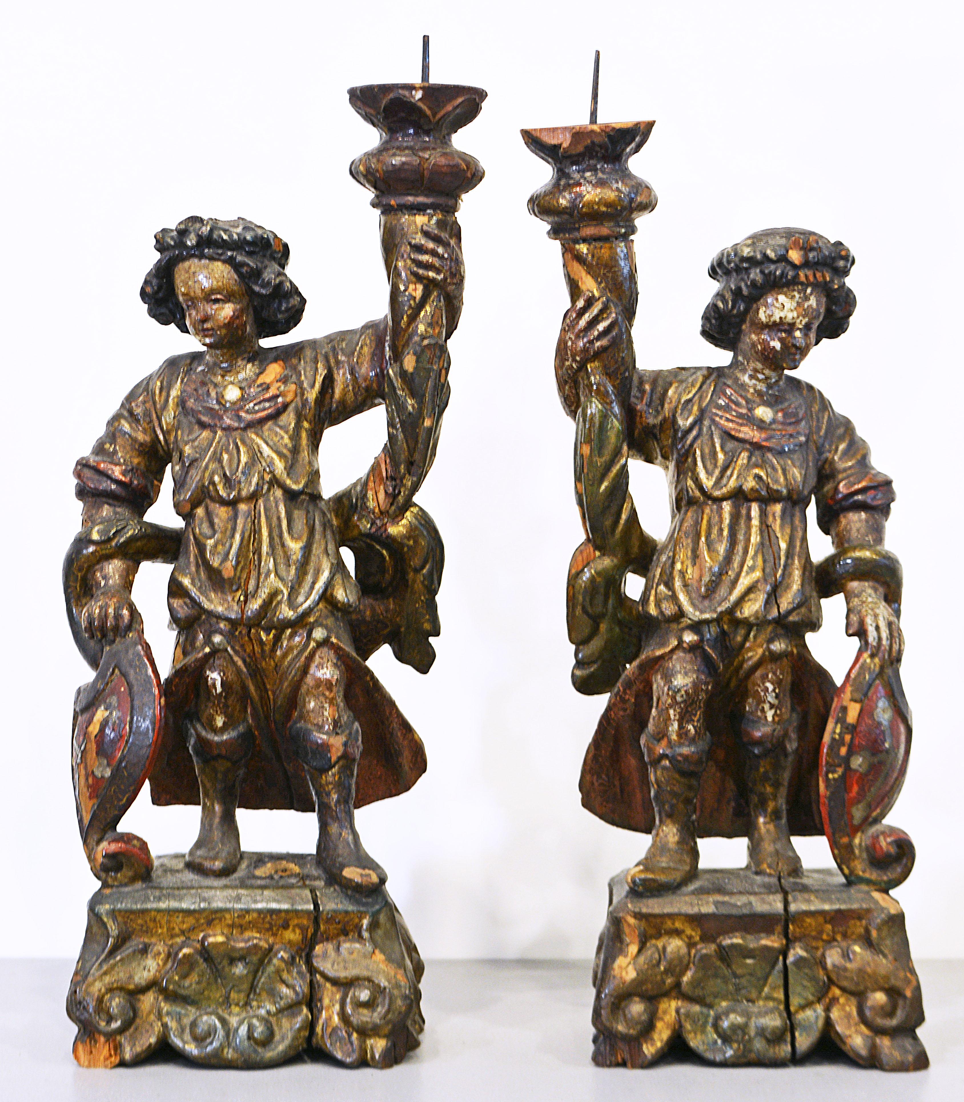 This rare pair of carved baroque torchiere saints, each standing contraposto wearing ornate billowing tunics and holding each a cornucopia shape pricket torchiere, raised carved square bases. Both saints are supporting shields decorated with the