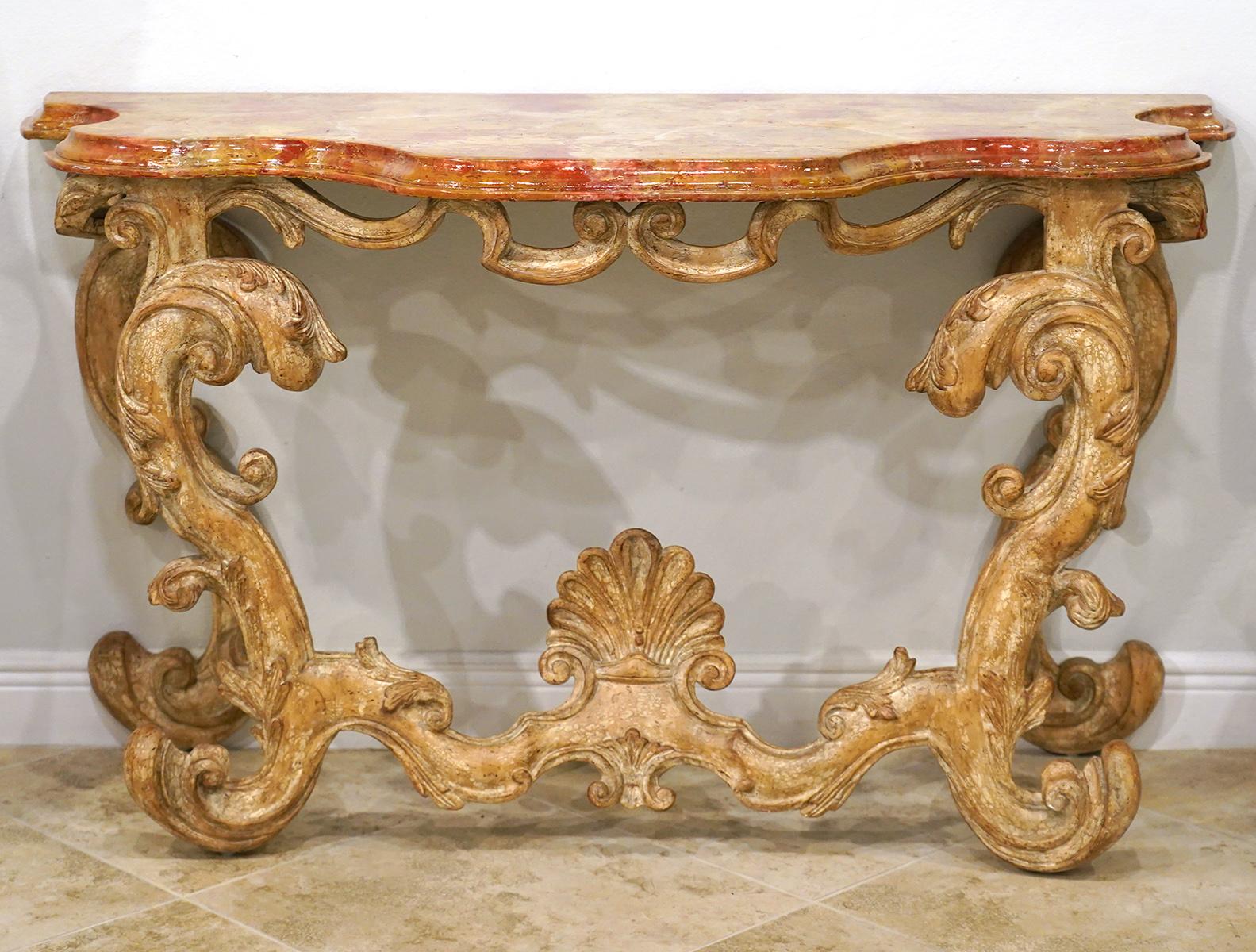 This pair of Italian Baroque style carved console tables feature marbleized shaped and molded tops above sumptuously carved frames in blonde wood with traces of paint. The front stretchers center elegant shell carvings.