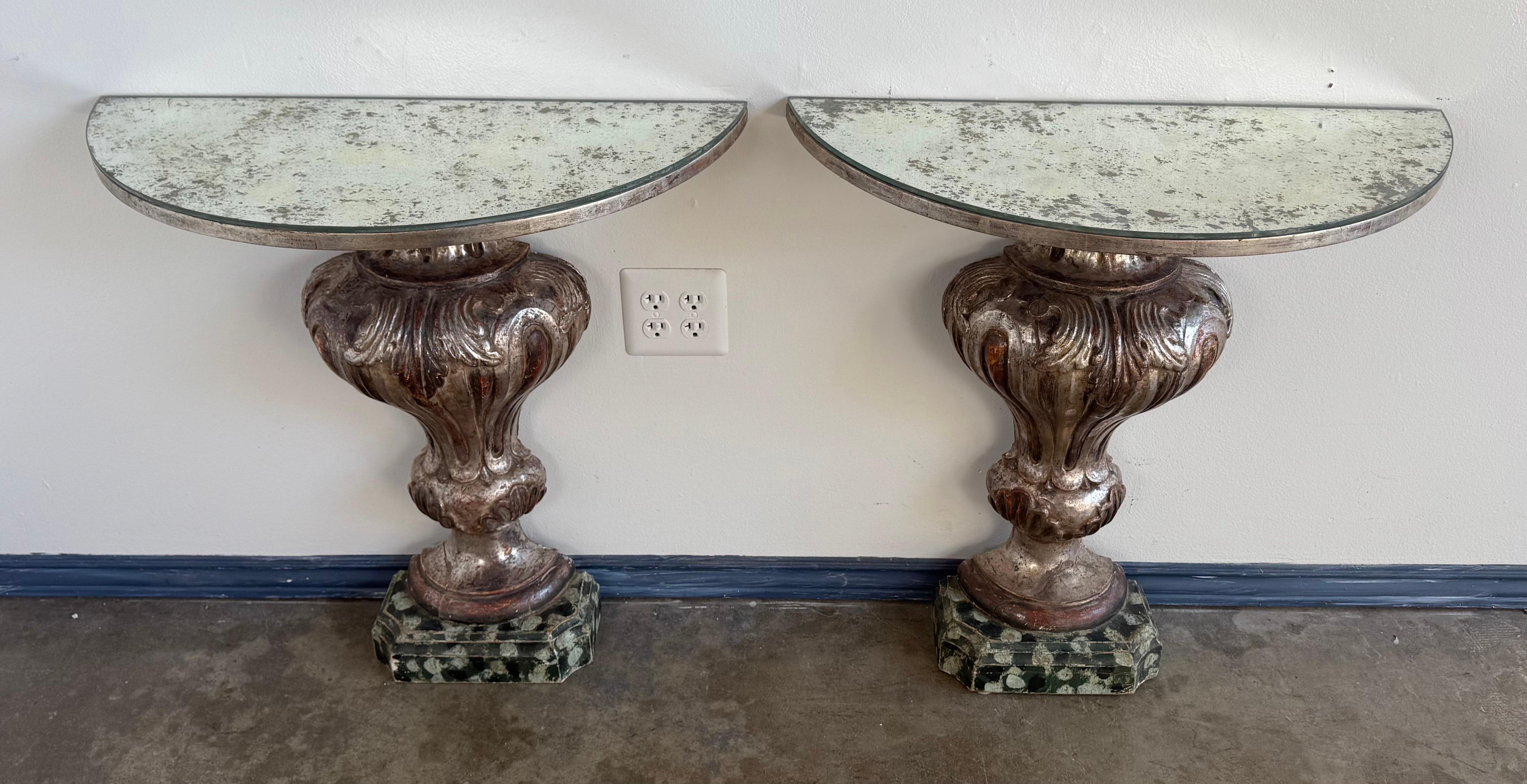 A dramatic and sculptural pair of Italian Baroque style wall-mounted console tables, richly finished in silver gilt with a beautifully aged patina. Each demi-lune console features a shaped mirrored top with a softly distressed surface, supported by