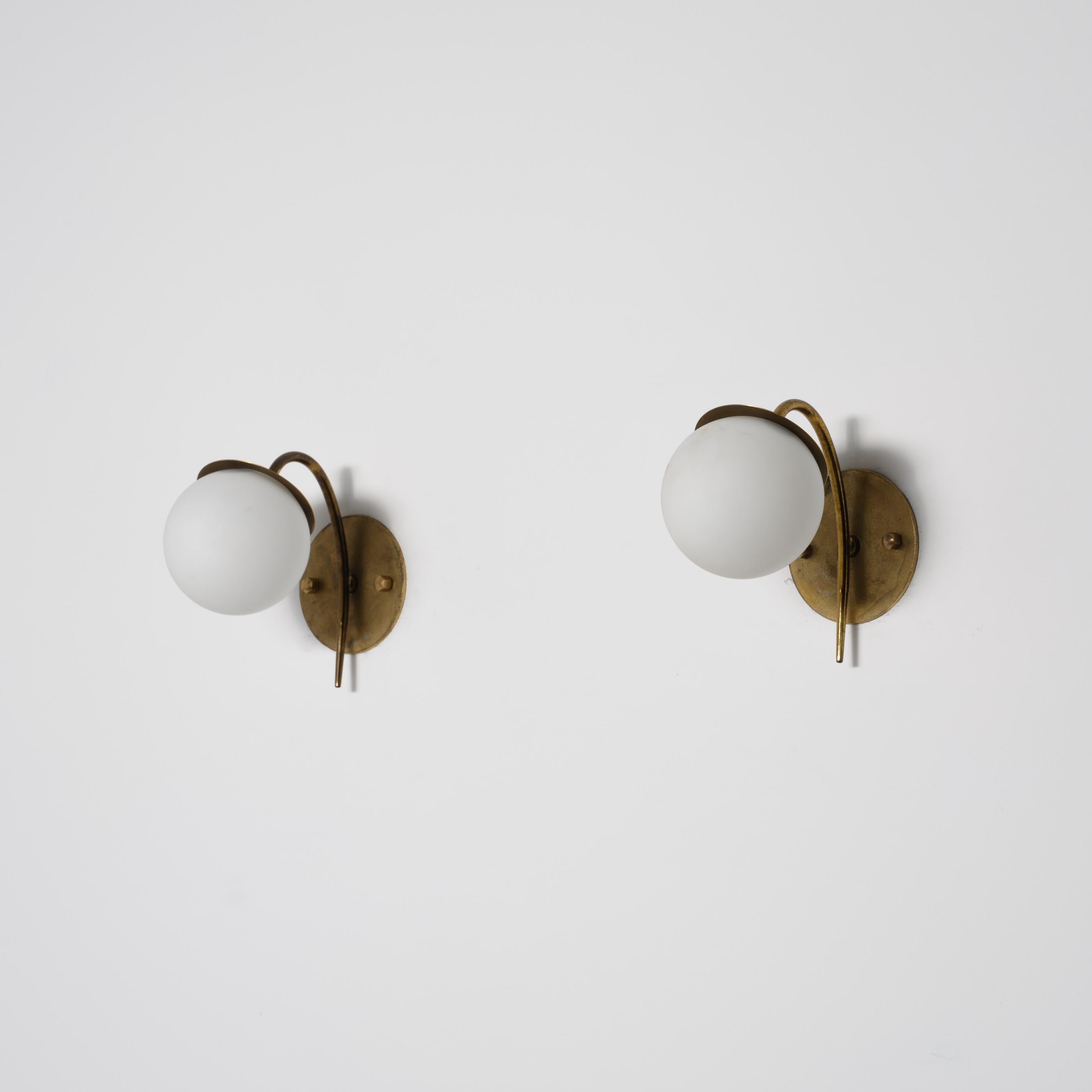Pair of Italian wall sconces from the 1950s, characterized by an elegant and sculptural brass structure with a naturally aged patina. The design features a curved arm that rises from an oval brass wall plate and gracefully supports a spherical
