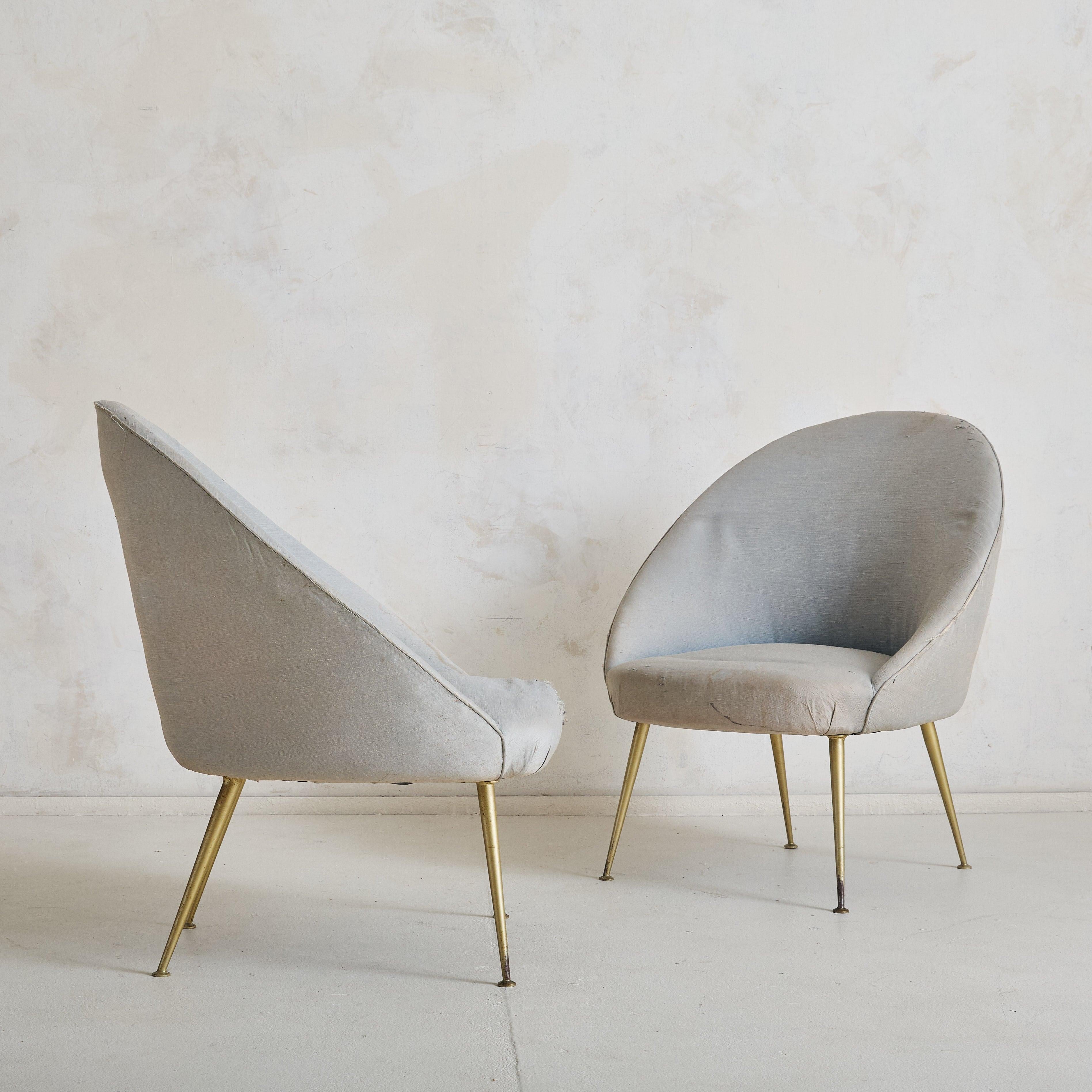 A pair of 1950s Italian lounge chairs, defined by their softly curved, capsule-inspired silhouettes. The minimalist frames are elevated by tapered, angled legs in solid brass. Reupholstery required; we have elected to sell these chairs in their