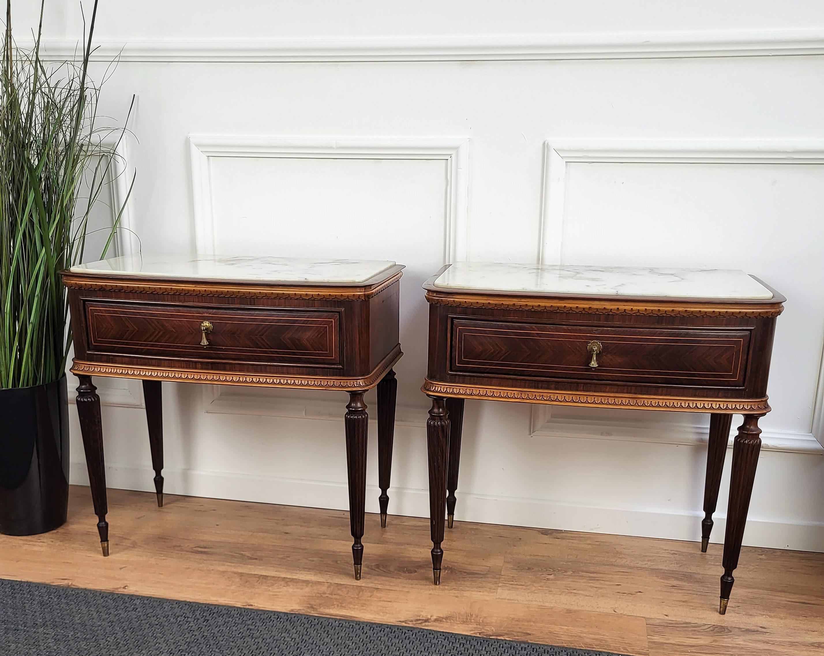 Very elegant and refined Italian 1950s neoclassical pair of night stands bed side tables with greatly carved detailed and decorated wooden structure, wood veneer decorated front drawer and white carrara marble top with brass details and fluted