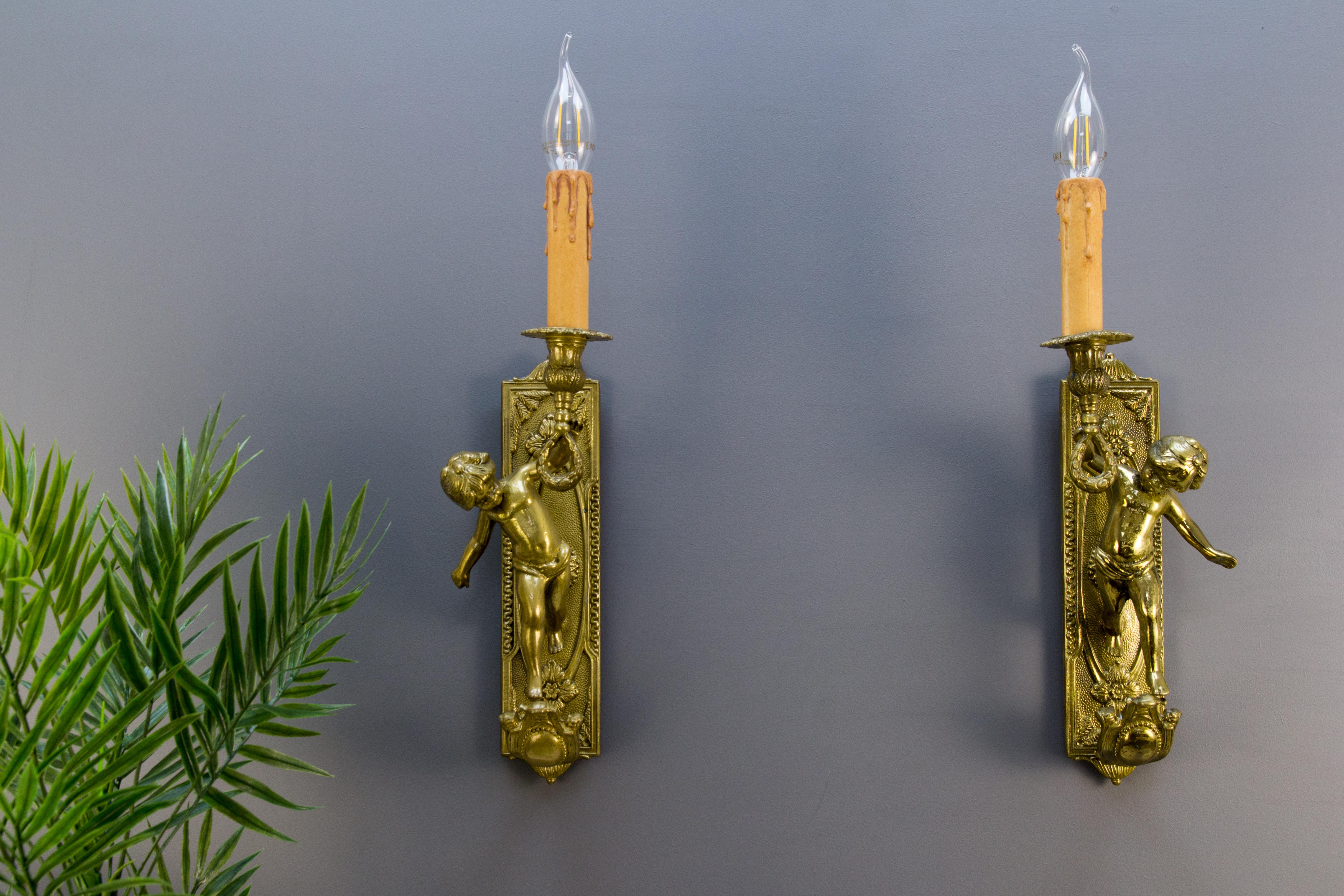 Pair of beautiful Italian electrified wall sconces, originally candlesticks made of brass and bronze in the 1950s. The wall panels are accented with neoclassical motifs on a textured surface. Cherub or Putti figure is holding up a torch that has a