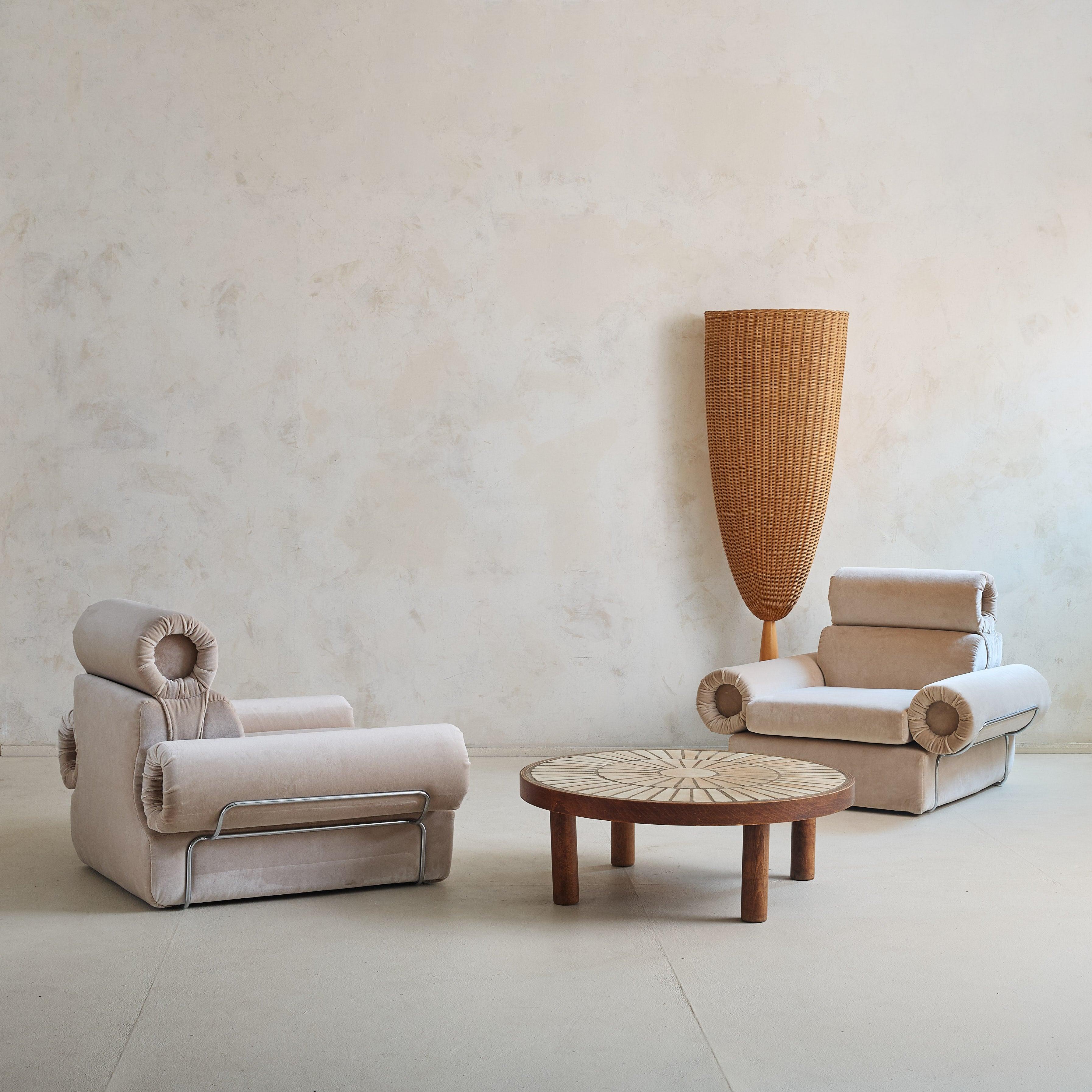 A pair of Italian lounge chairs from the 1970s, embracing bold geometric shapes and plush forms. The cushions are complemented by rolled, cylindrical arm and backrests, balancing a playful yet structured design. Freshly reupholstered in pale fawn