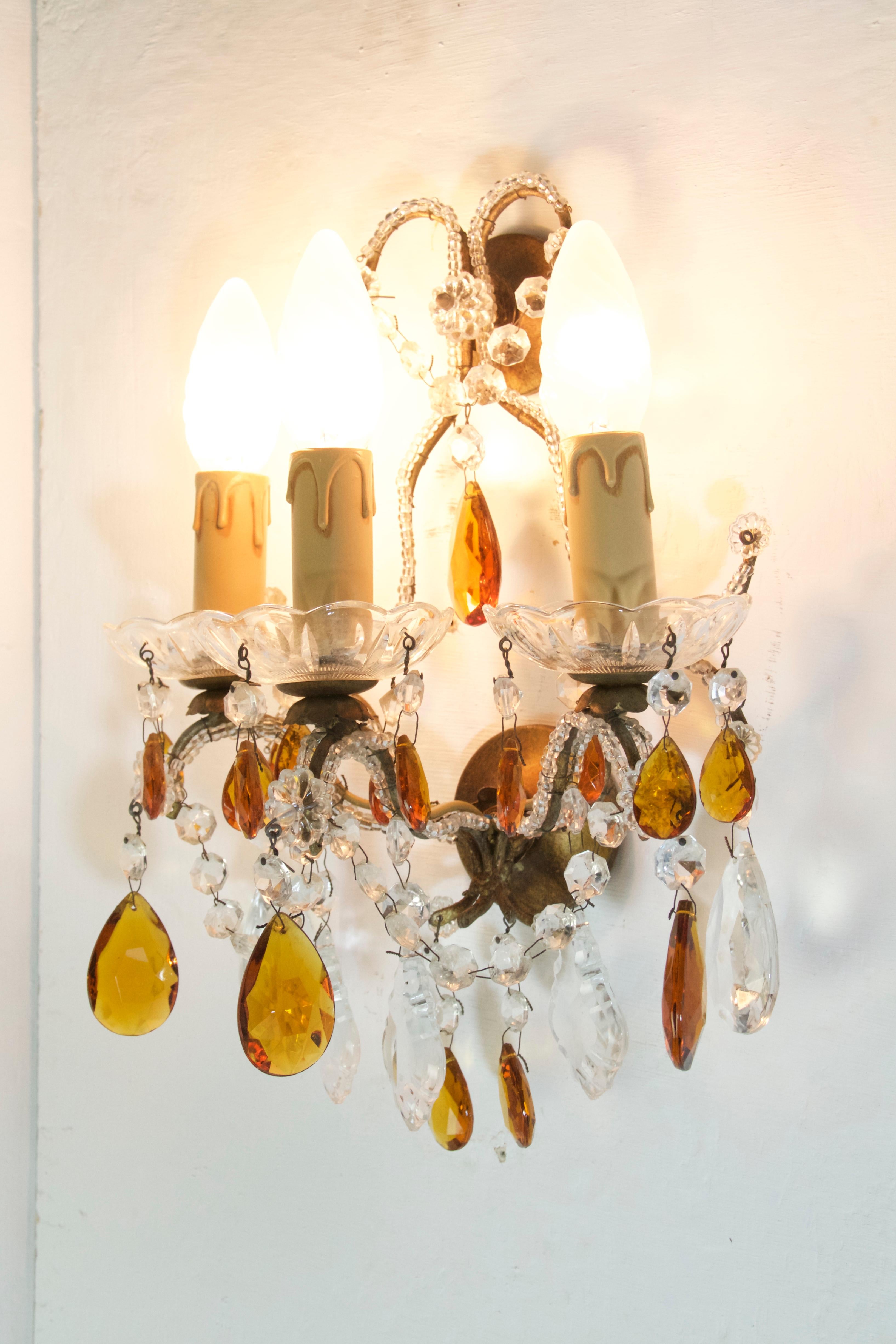 Two sets with two pairs in each set of wall sconces in the manner of Maison Baguès. Each sconce has three candle style bulb holders and is decorated with clear and amber colored crystals and glass. The sconces are made from iron and painted in gold