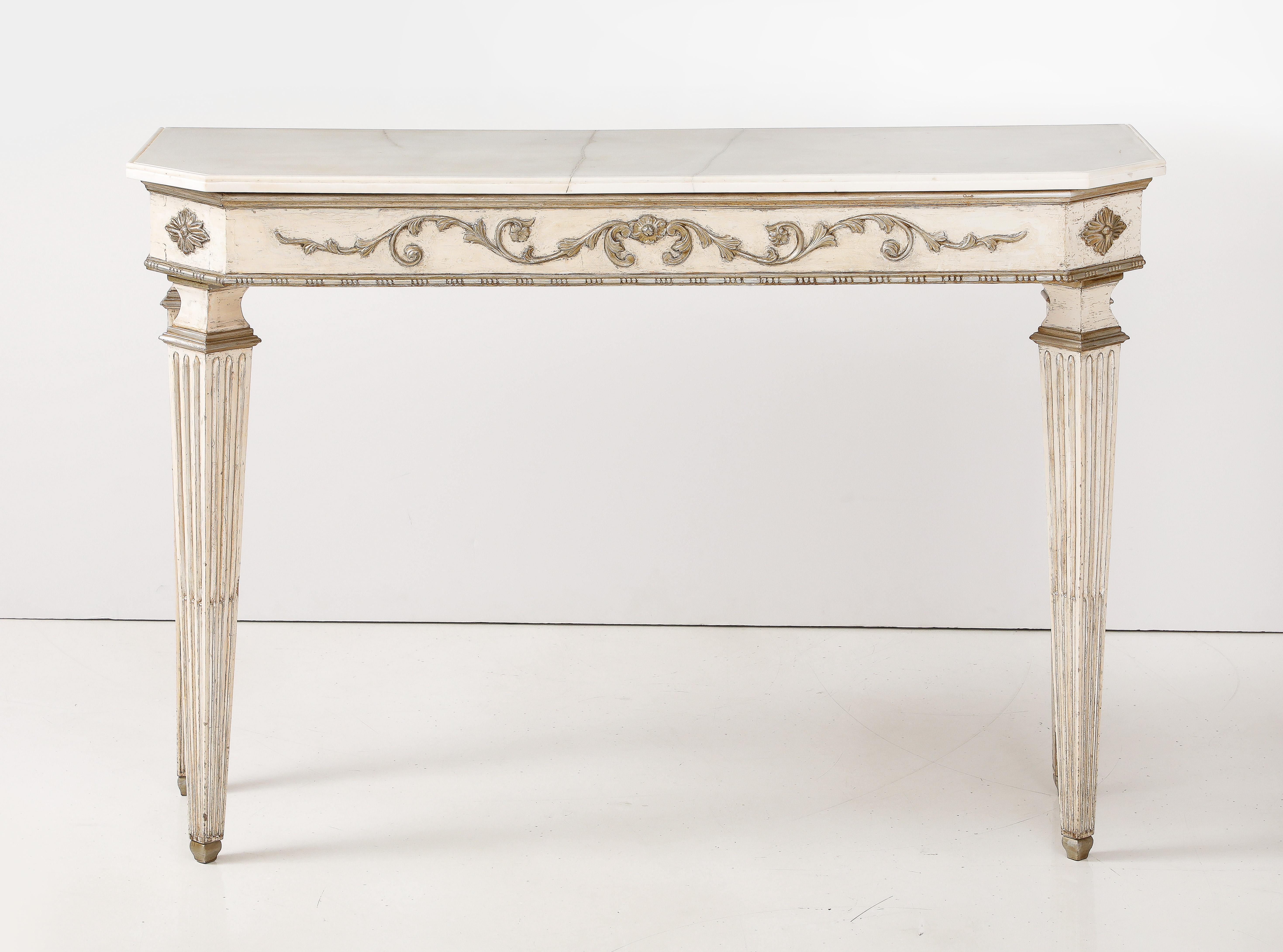 Pair of Italian Directoire console tables; the carved, fluted and tapered legs support an apron with silvered motif of rosettes and fanciful carving on all sides.  The whole supports the original Carrara marble shaped tops following the design of