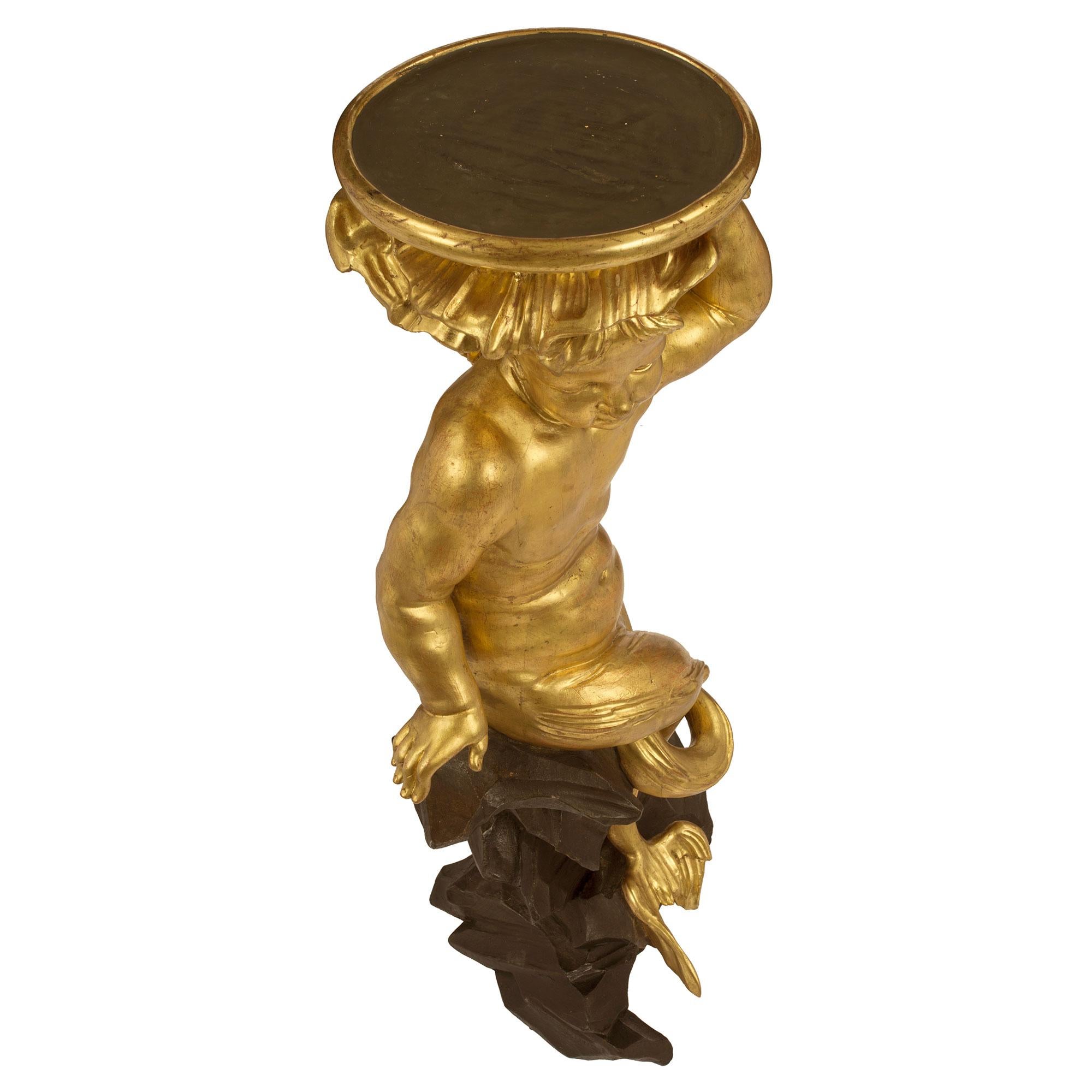 A sensation and most elegant pair of Italian early 18th century Roman giltwood and black polychrome pedestals. The pair are raised by a rock like grotto base in a dark polychrome below a seated merchild. Each charming life size gilded merchild has a