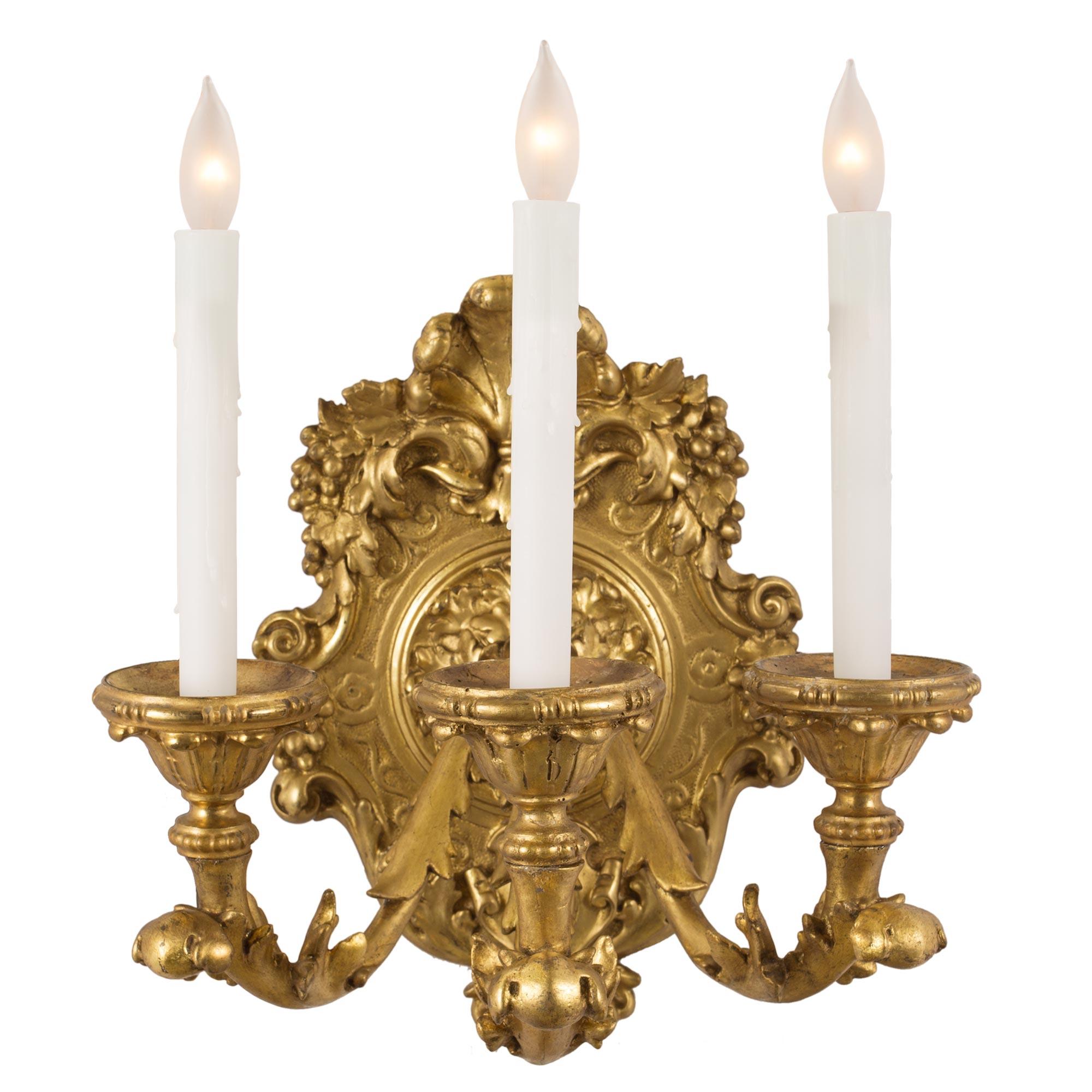 A pair of Italian early 19th century Louis XV st. three-arm giltwood sconces. The sconces with an oval back plate are decorated with richly carved acanthus leaves amidst scrolls. At the top is a central feather spray flanked by grape clusters and