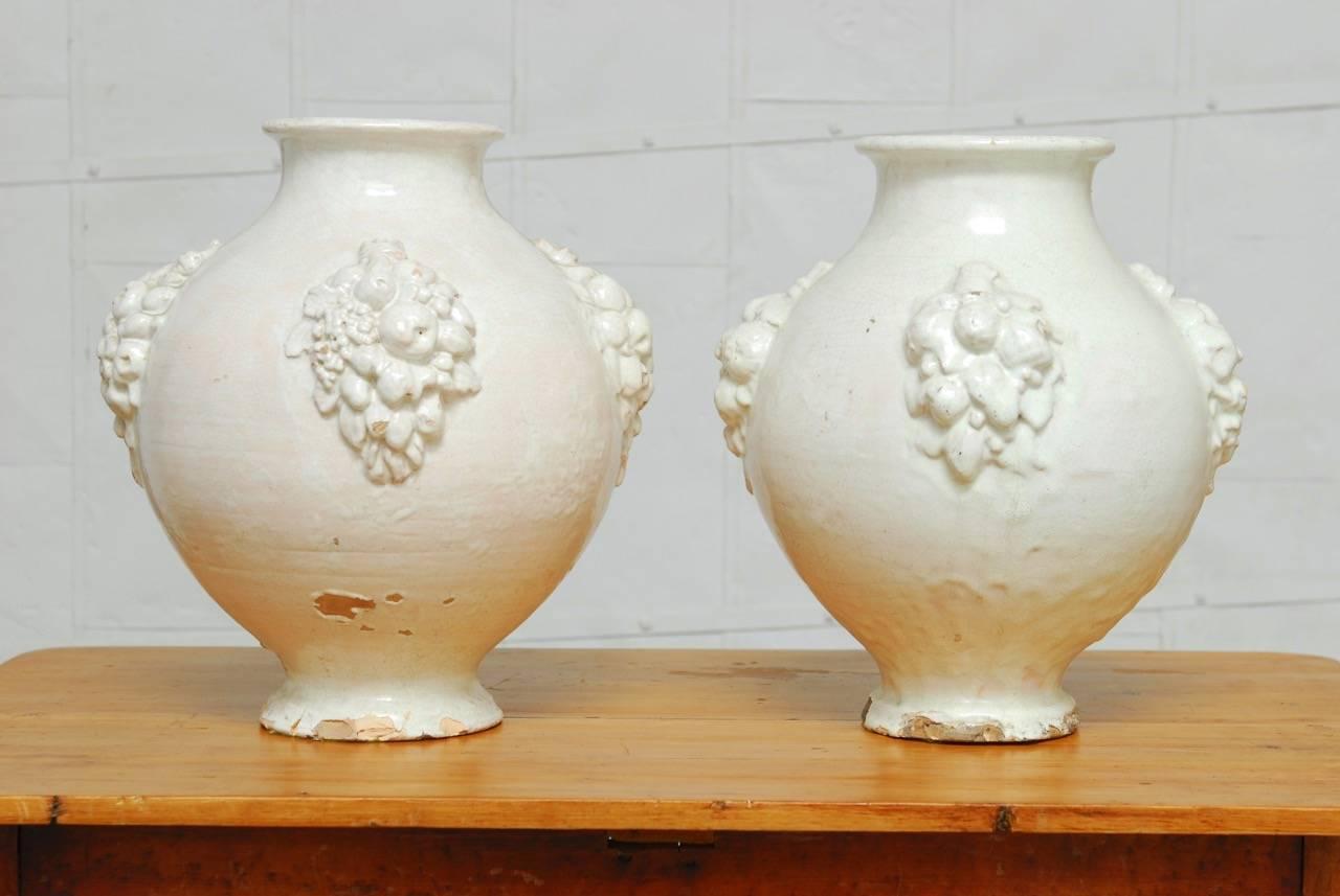 Pair of rustic Italian earthenware pottery jars with a bulbous urn form body and fruit clusters cascading down the tapered base. Featuring a thick cream ware glazing that has a rich craquelure finish. Est. circa 1900, these jars have chips and