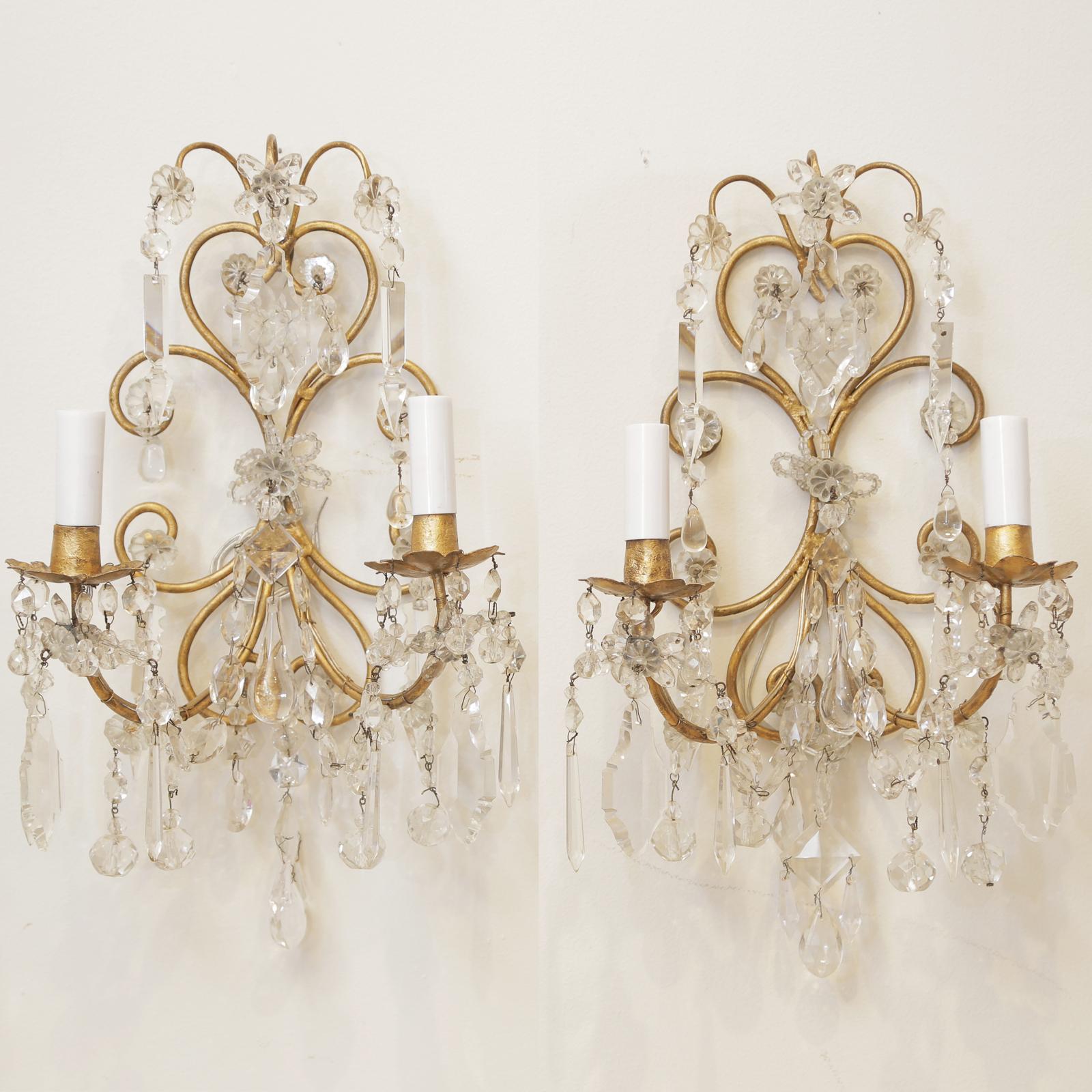 Pair of sconces, each having a backplate of scrolling gilt metal, adorned with faceted crystal prisms, drops, and pendants; floral rosettes petals pay homage to the styles of Maison Bagues, two C-scroll candlearms extend from the backplate and