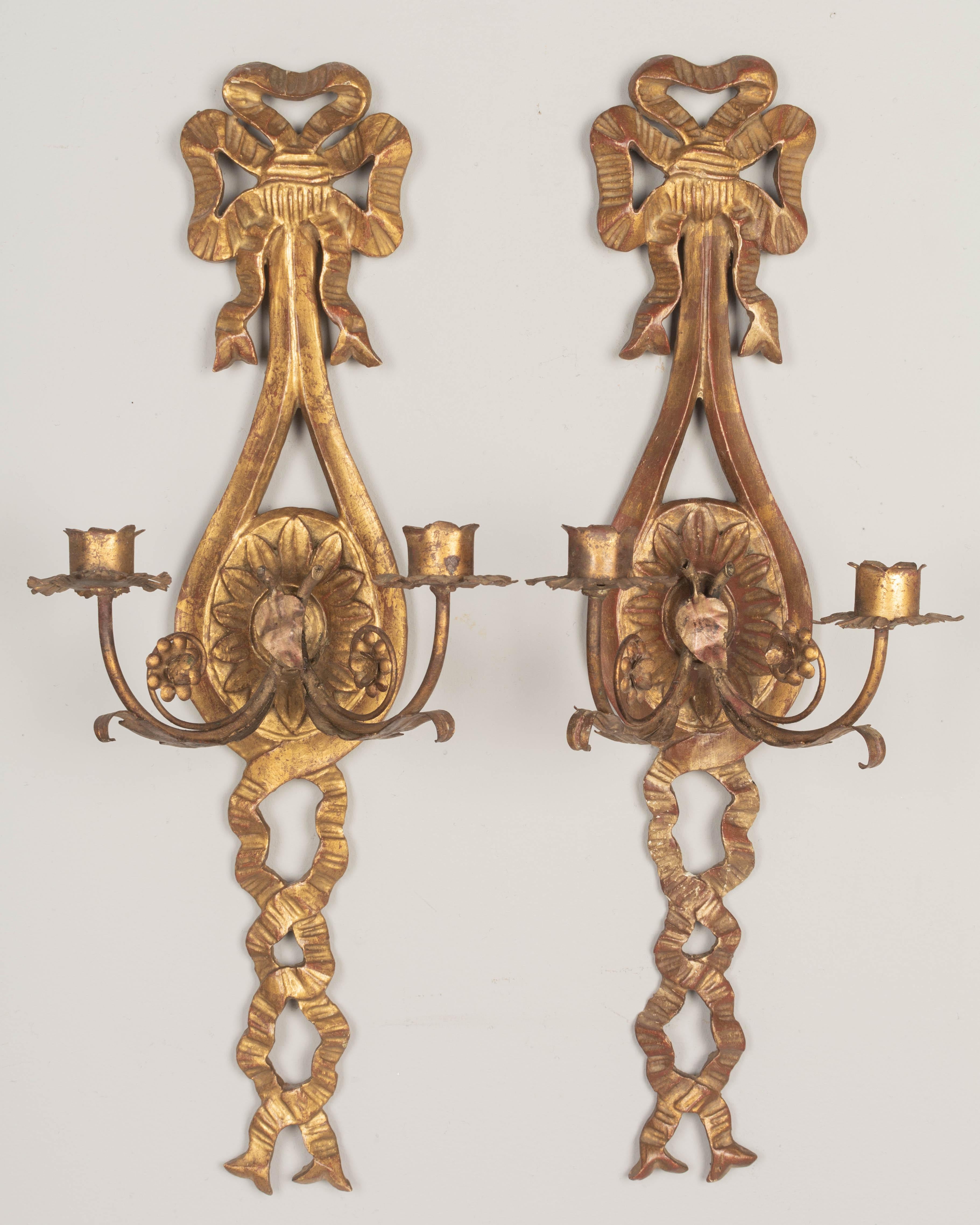 A pair of Italian two-light gilded wood candle sconces, each with carved bow and ribbons. In good condition with warm gilt patina. Two scrolling metal candle arms with tôle details. One arm restored. Not wired, but may be electrified. Circa