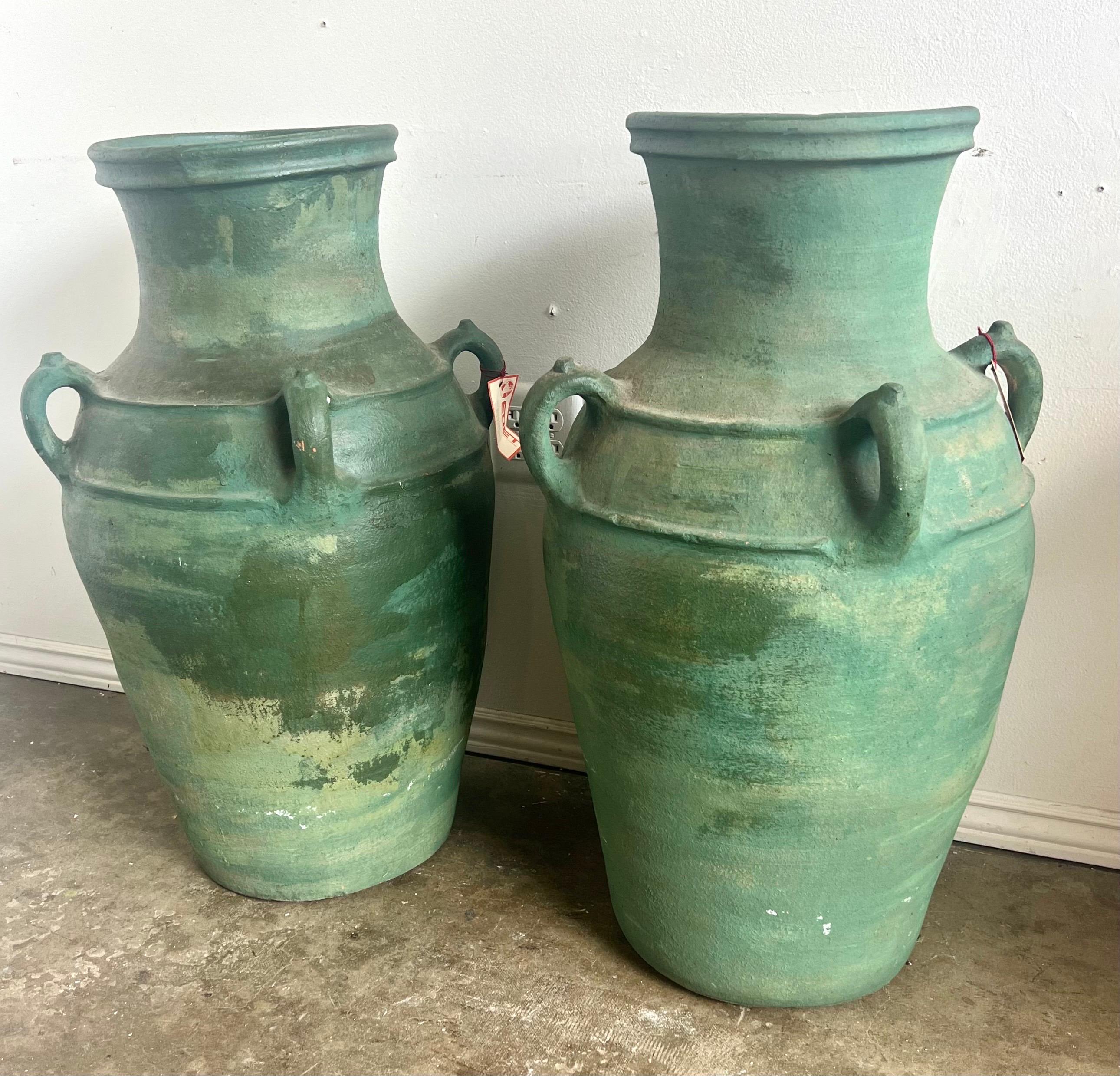 Paire de grandes urnes en céramique, chacune avec un corps robuste et arrondi et un col étroit et évasé.  Les urnes sont recouvertes d'une glaçure verte vieillie qui leur donne un aspect antique.  Les deux urnes sont équipées d'une double poignée
